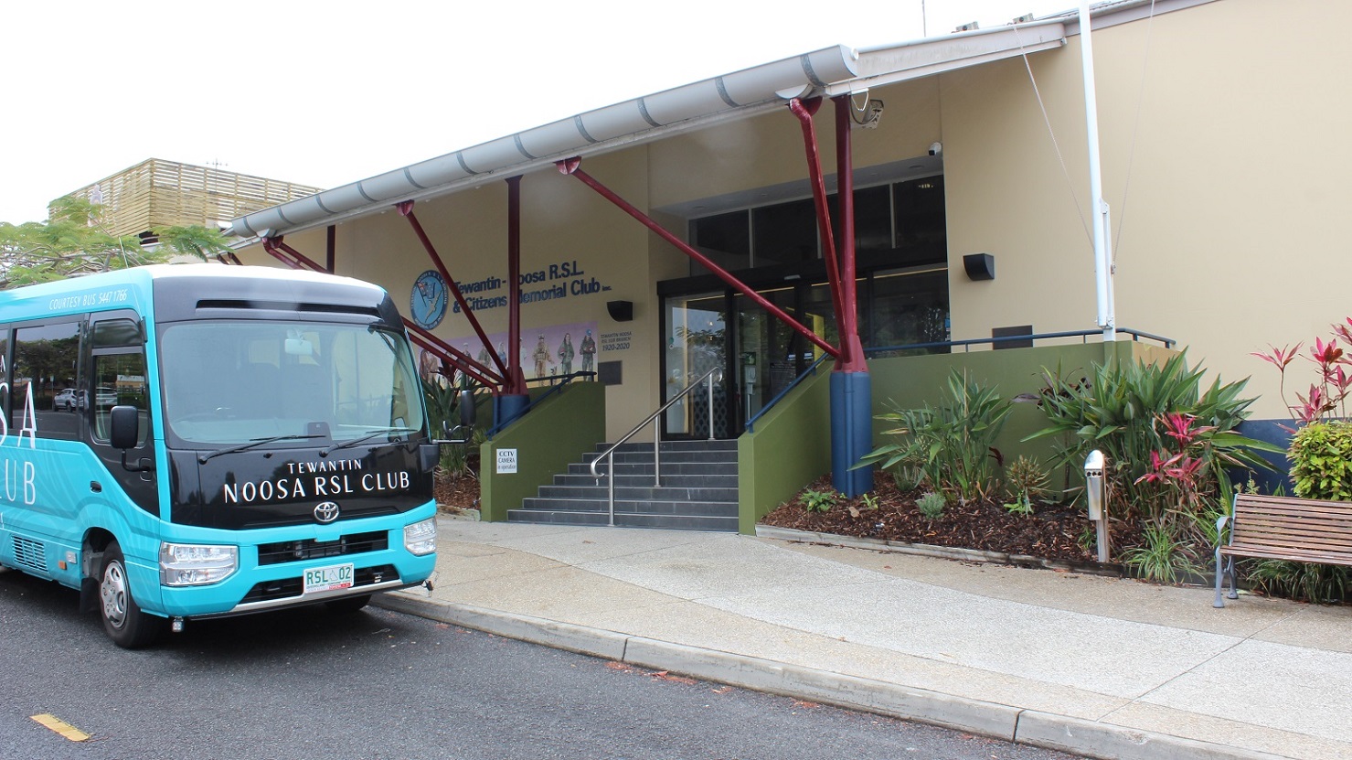 Another view of the previous image focuses on the glass sliding door and black tiled steps under the red/blue pillar-supported exterior. A steel rail in the centre divides the stepway into two halves bordered by green-painted half-walls with rails on both sides. Plants are in triangle forms by a seat on the right facing the sidewalk and parking lot. The bus is packed in front.