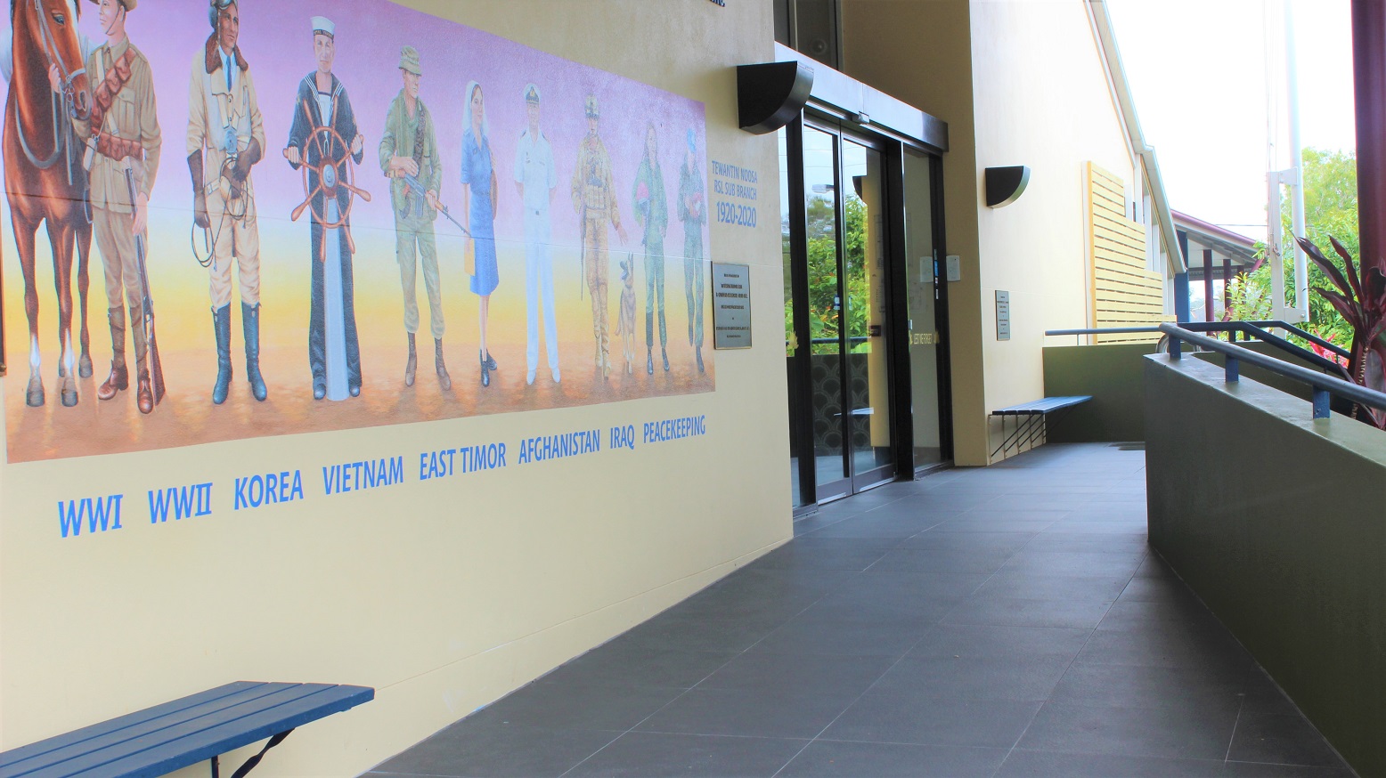 A closer view of the yellow wall with military artwork, engraved inscriptions, and blue wall seats on both sides on a grey-tiled walkway flanked by green-painted half-walls with railings.