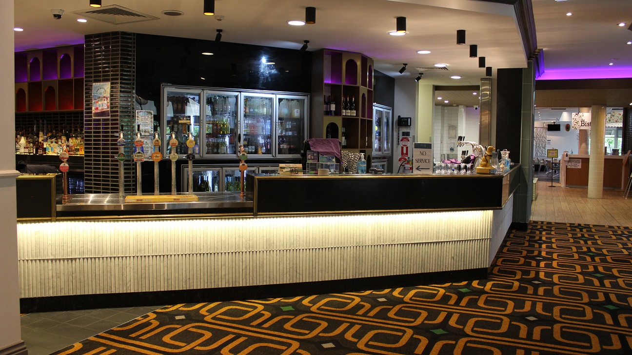 A view of the cafe's counter, designed in black and white and has two sets of broad fridges packed with drinks separated by a wine rack behind the counter. On the left by a pillar is another wine rack packed with wines. On the counter are a screen, infographic, pos, sanitizers, and other items on the right. Beers on tap steel covers a lower spot on the counter. The floor is covered in an army theme. The walkway leads to the bistro-themed restaurant.