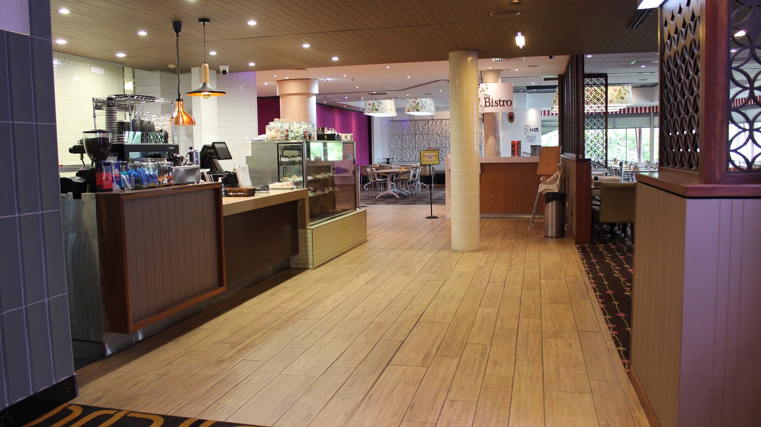 The Bistro restaurant is accessed through a beige-covered floor, with a brown designed counter on the left having coffee packs,a cabinet with cups, a checking screen, and a lit snacks showing cabinet. On the left is a wide entrance with walled space having a rubbish bin and seats. Two white pillars face the bistro tag on a flowering white lamp cover and the direction board on the entry to the purple and white wall dining room with seats and tables on the right.