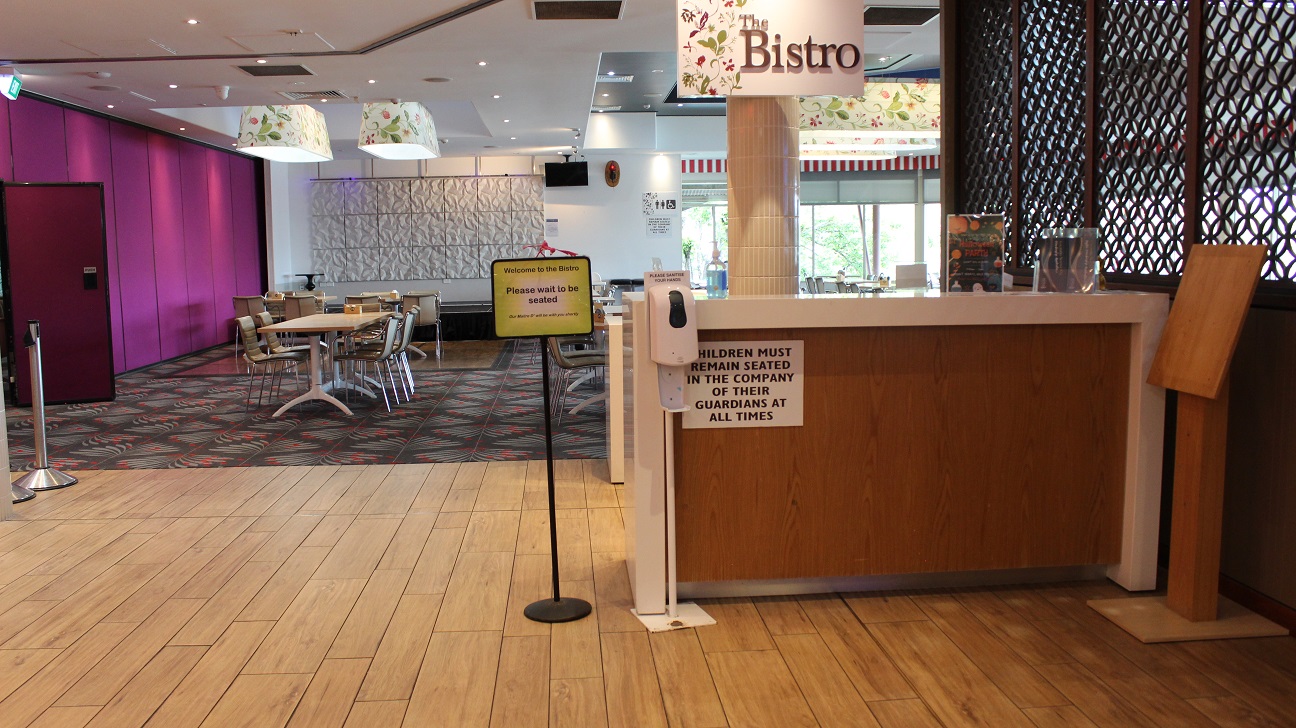 A close-up view of the Bistro restaurant. On the right, the Bistro is written on a lamp cover by a pillar on a beige-tiled floor with a brown wooden accented counter. On the desk is an infographic, a menu, and hand sanitizer. Direction board, a sanitizer dispenser, and a wooden board are seen by the counter. The hallway leads to a purple and white marble wall by the seating area with sets of four brown tables arranged around a table on grey and red tiles.