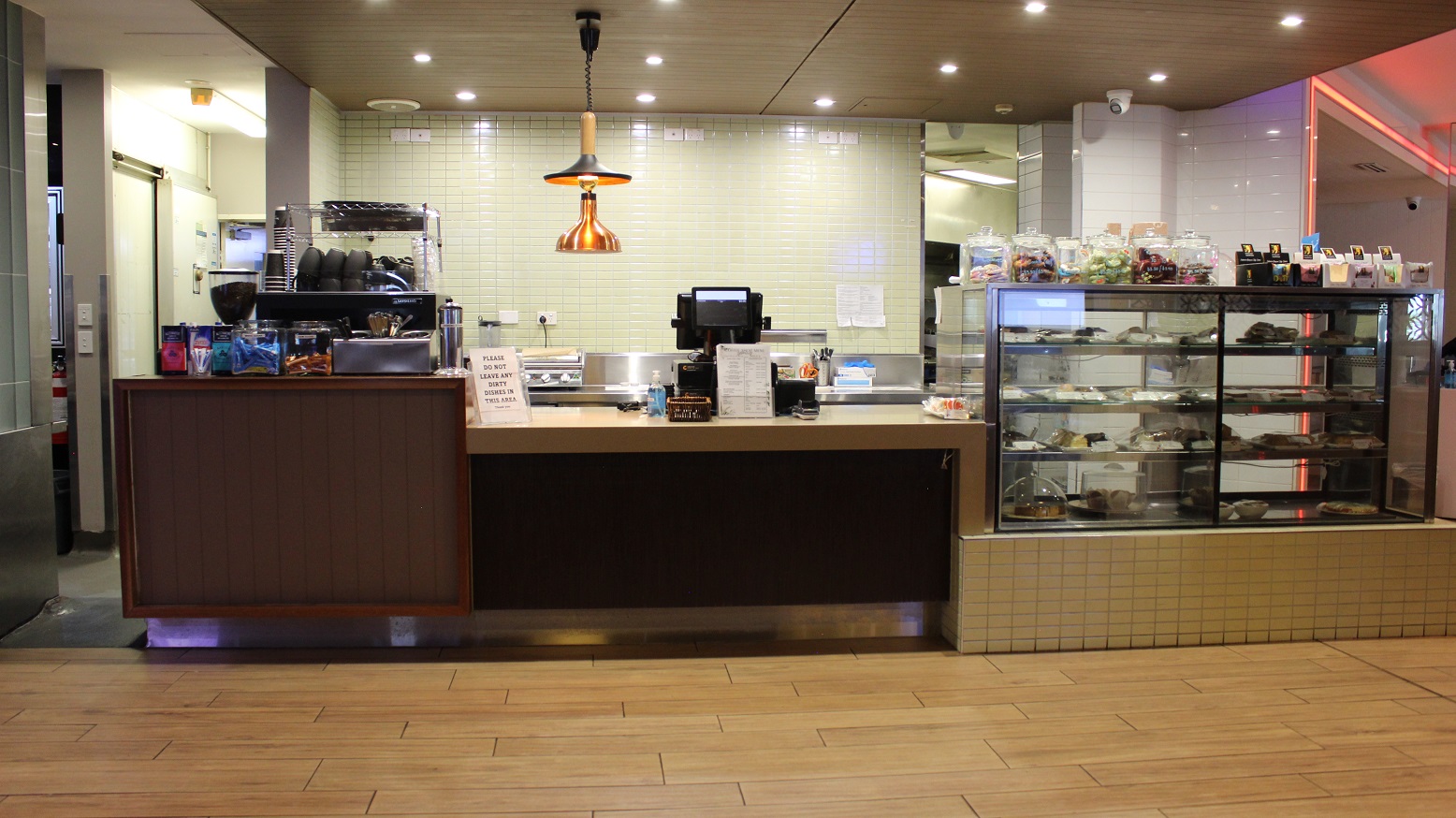 Another view of the café counter with a brown accents on the beige tiled walkway. The desk to the left have coffee packets, a large glass, a spoon and fork vessel, a flask, and a coffee cabinet with cups. The right, a food display shelf with cakes and snacks has snack jars and packers neatly organized on it. The checking screen, infographics, and sanitizers are on the middle. Behind are silver kitchen wares on a beige wall with an infographic and a mirror to the right.