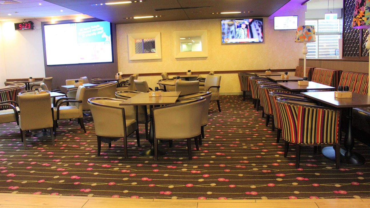 A brightly lit dining room with rows of chairs and tables. On the left, there are a few multicolored seats set horizontally against the wall, along with a large and small screen. Two heat stands are visible. In the centre, two rows of brown chairs and tables lead to a wall with a yellow frame pattern. The last row of multicolored chairs leads to the wall with a big screen. On the left and a timer. The floor is coated in a dark grey with a pink and yellow motif.
