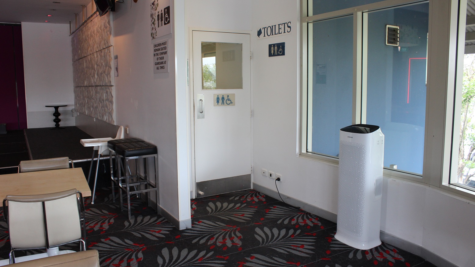 Behind a white marble wall with an infographic by the purple wall overlooking a space with a white highchair and two black high stools leading to the toilet. A white door with an infographic and a toilet tag on a grey and red covered floor surface, flanked by a white wall and a glass window on the left. A purifier can be seen standing by the wall on the right side.