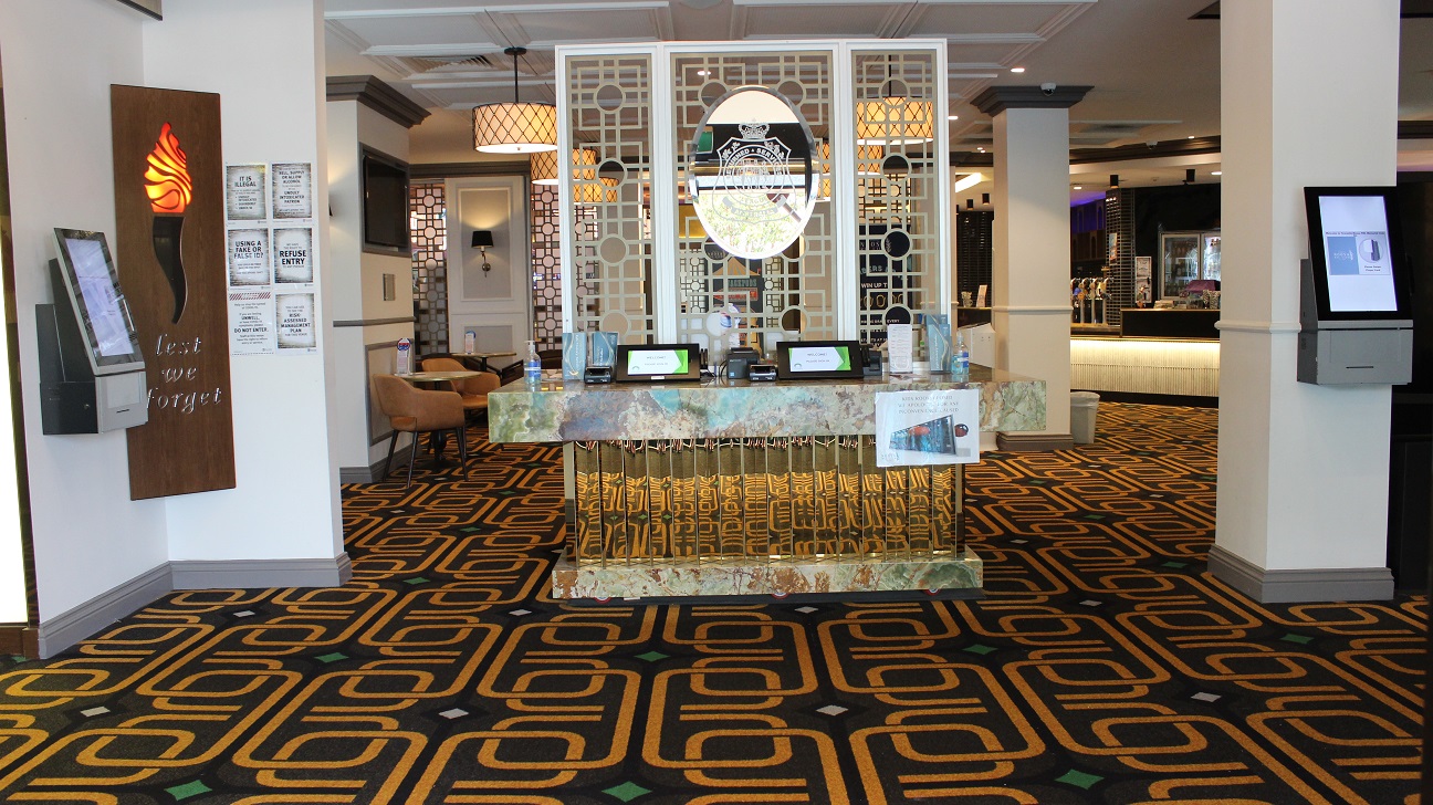 A table with a military emblem made of marble (white, brown, and vivid gold) stands in the centre of a foyer covered in brown and vibrant gold carpet and accents, barricading the seating space. Brown chairs, tables and a TV are on the left wall. Two pillars are visible, one with a check-in screen and the other with a rubbish bin. Ticket dispensers, infographics, and sanitizers, among other things, are on the table. At the far end, is the café.