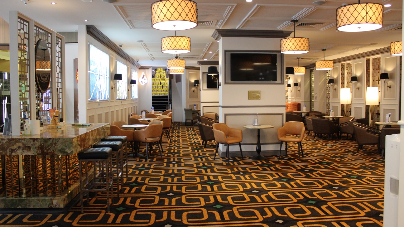 The dining area behind a table with the military motif emblem, and three stools resting on it. A napkin, ornaments, and other items are on the table. The well-lit area features brown chairs placed in rows around each table on a brown bright gold carpet. Chairs and tables lean against the two pillars, with TVs in the centre. At the other end, there are two orange seats. Every table has a menu in glass decor. The walls are white with brown accents and bright gold accents.
