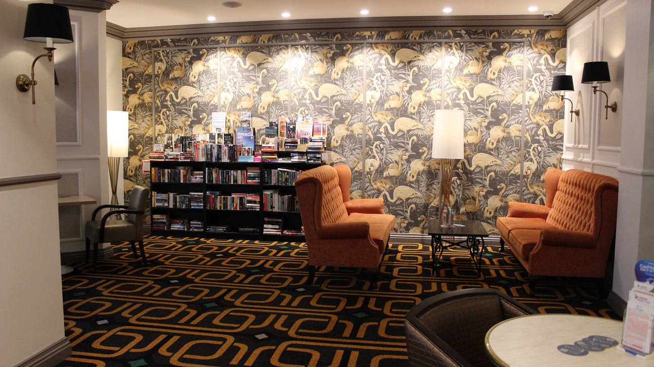 A view of the brown and yellow flamingo-themed wall, with a bookshelf to the left and two white-covered lamp fixtures. On the left are two sets of two cozy orange seats with a dark brown table in the centre. The walls are white, with a dark brown accent, and the floor covering is brown and gold.
