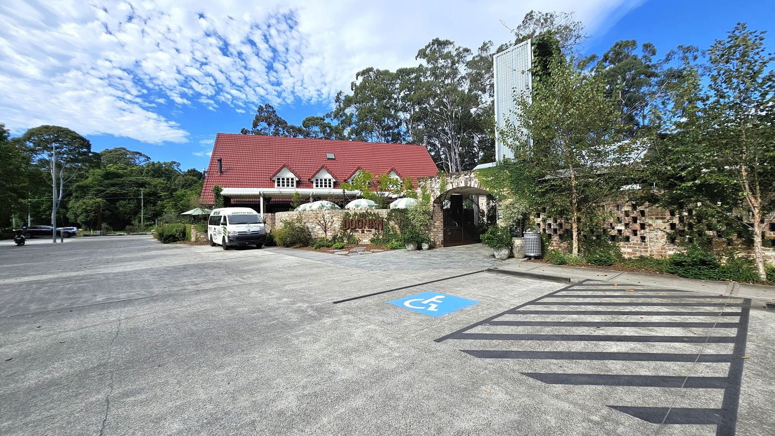 A building with a red gabled roof and multiple dormer windows is in the centre. In front, a white van is parked near a brick wall with a sign. Several white umbrellas are visible behind the wall. On the right, there is a patterned brick wall with greenery and trees. A blue disabled parking space is marked on the concrete ground in the foreground. Tall trees are in the background, and a few vehicles are on the left side of the parking area.