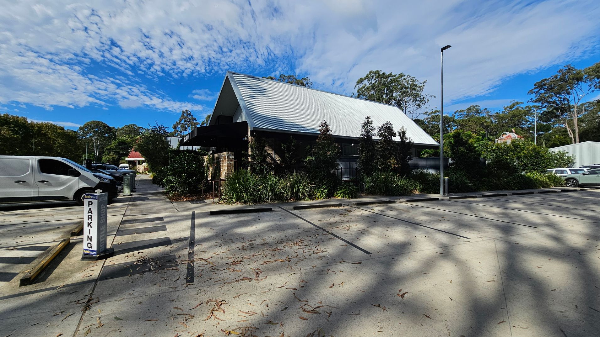 A parking area with a sign marked "PARKING" on the left. Several vehicles, including a white van, are parked. In the centre, a building with a steep, corrugated metal roof is surrounded by greenery. The right side shows more parking spaces and a tall streetlamp. The ground is concrete with scattered leaves and shadows from nearby trees.