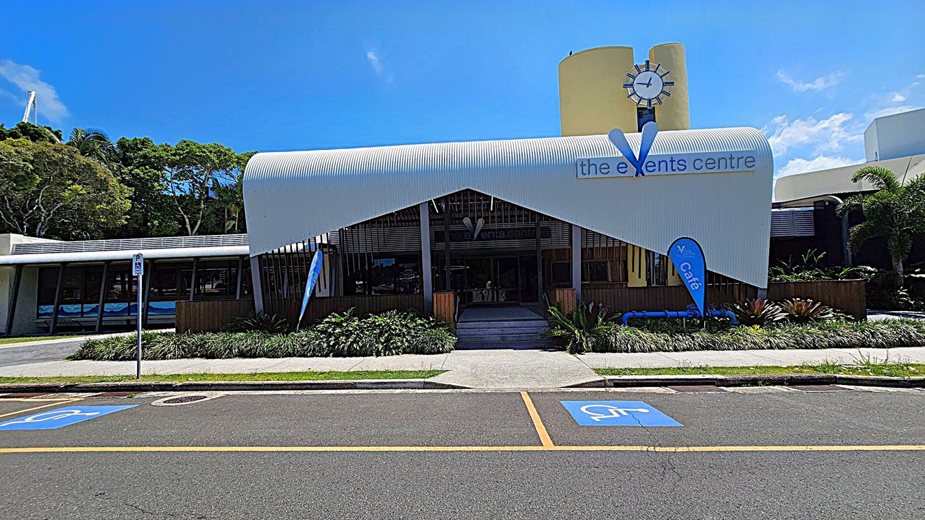 A modern building with a curved white roof and a sign reading "the events centre" on the front. A clock is mounted on a yellow cylindrical structure above. Blue flags with "café" are displayed near the entrance. The foreground features a road with marked disabled parking spaces and a pedestrian path lined with greenery. Trees are visible to the left, and palm trees are on the right.