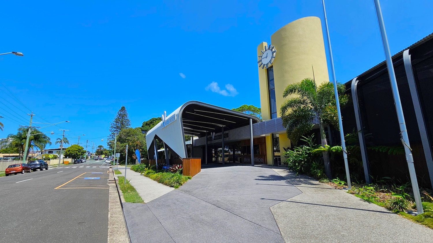 A modern building with a curved entrance and a large clock on a yellow cylindrical tower on the right. The foreground features a wide pavement with greenery along the edge. On the left, a street lined with parked cars and palm trees extends into the distance. The building has a sleek, contemporary design with a mix of metal and glass elements.