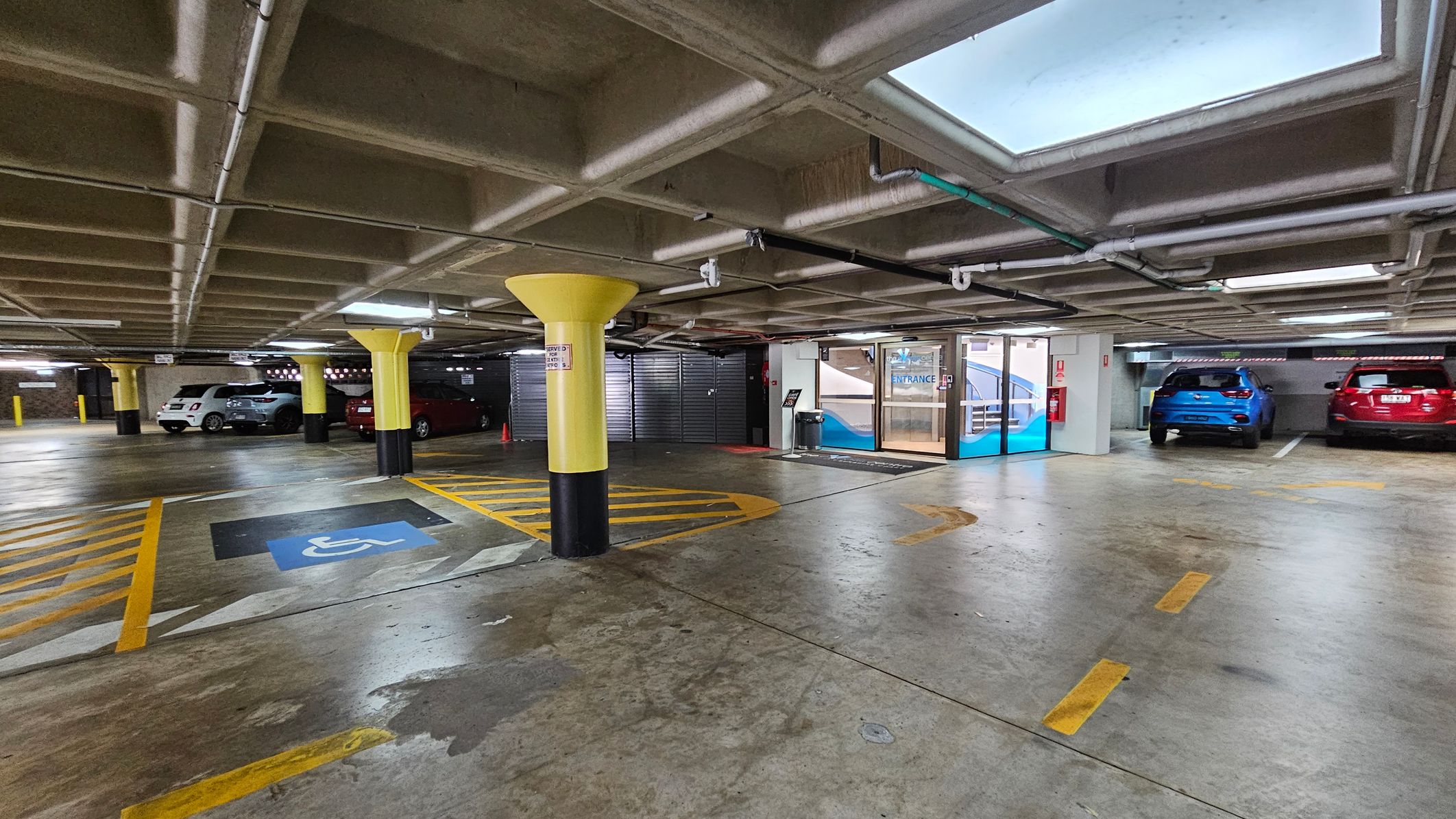 Underground car park with a concrete floor and ceiling. On the left, several cars are parked, including a white and a silver vehicle. In the centre, a yellow column stands near a marked disabled parking space with blue and yellow lines. On the right, a blue and a red car are parked. The background features a glass entrance door with a sign above it. The ceiling has visible pipes and rectangular skylights.