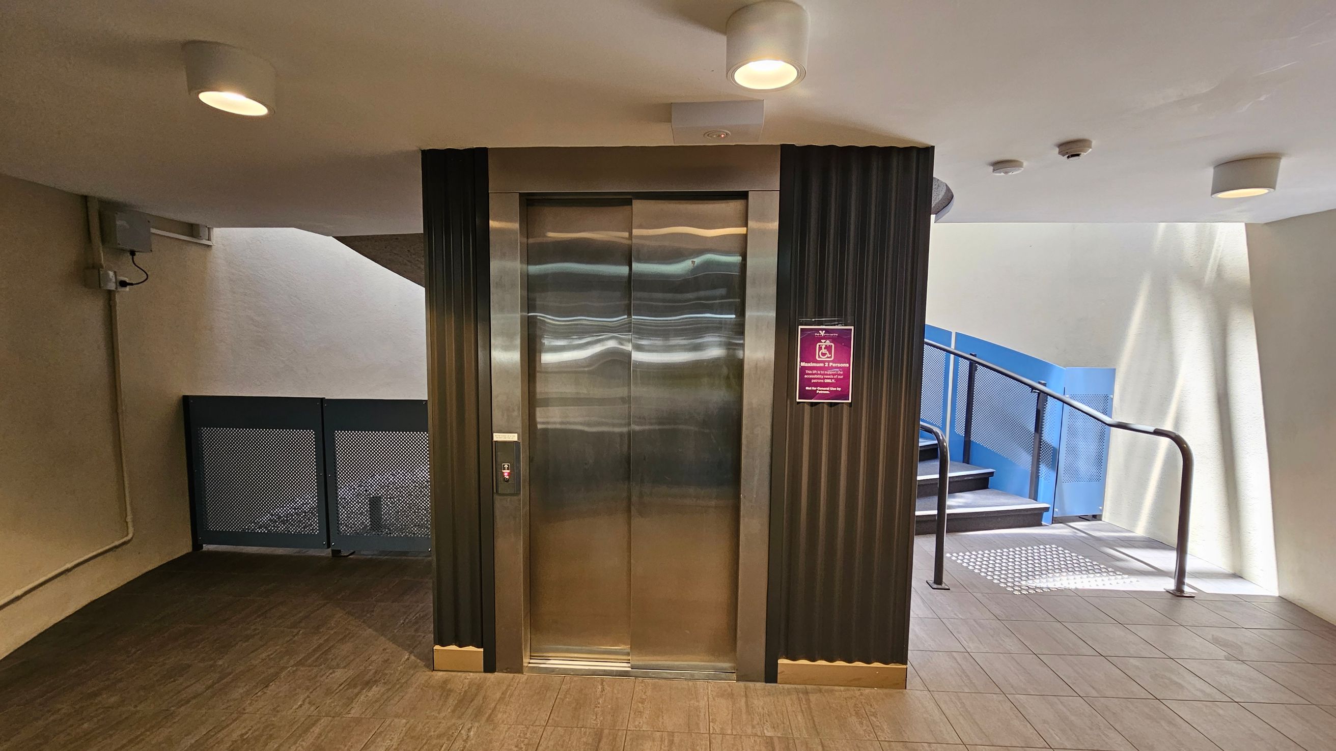 A modern lift with shiny metallic doors is centred in the image, flanked by dark vertical panels. To the right, a staircase with a blue railing ascends, with a textured mat at the base. On the left, a wall-mounted electrical box and a vent are visible. The floor is tiled in a light brown shade, and the ceiling is white with recessed lighting fixtures. A sign with a pink background is mounted next to the lift.