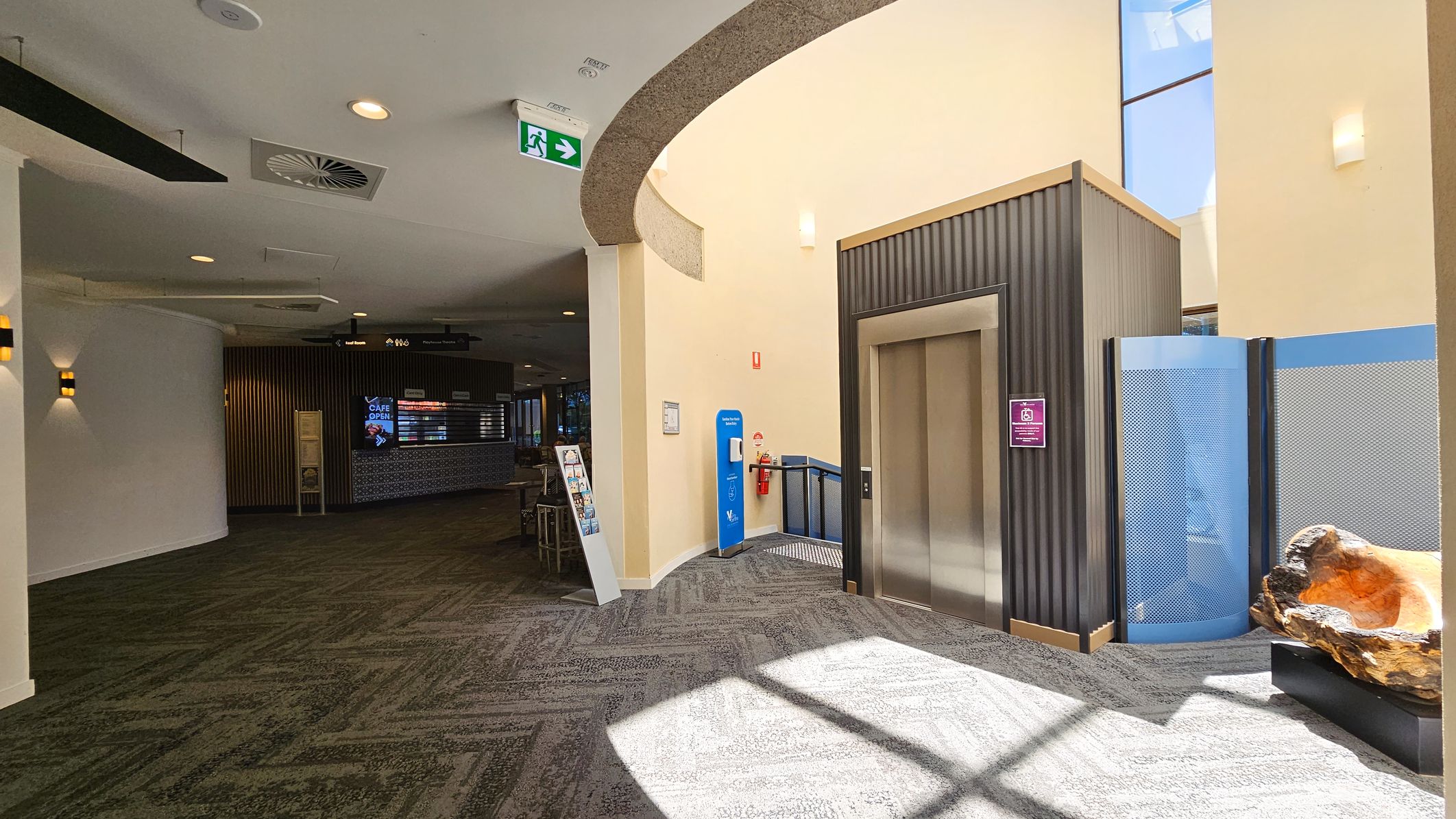 On the left, a curved wall with wall-mounted lights and a green carpeted floor. In the middle, an elevator with a metallic door, a blue hand sanitiser station, and a fire extinguisher. On the right, a decorative wooden sculpture on a black stand and a blue partition. The ceiling is light-coloured with recessed lighting and ventilation. An exit sign is visible above. In the background, a café area with a digital menu board.