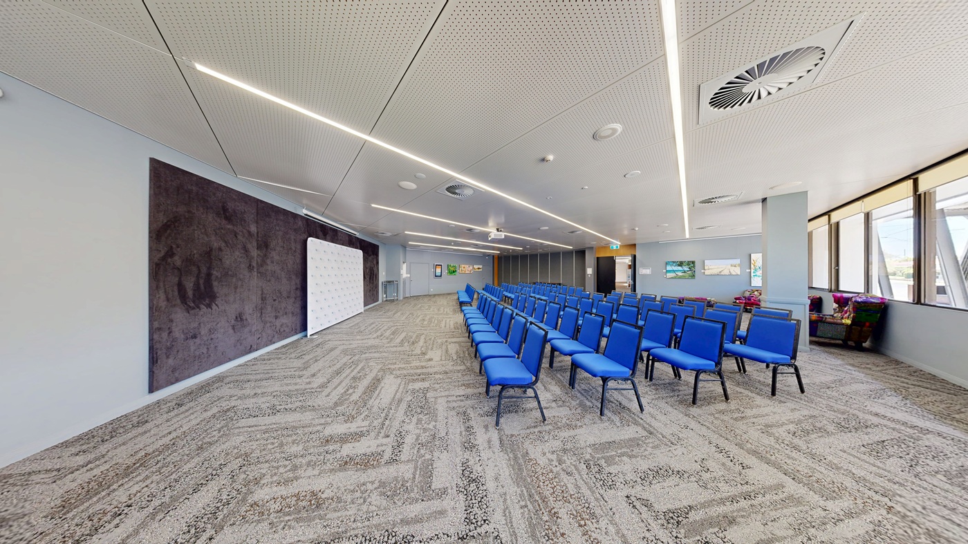 A conference room with rows of blue chairs arranged in the centre. On the left, there's a large dark panel on the wall and a white board. The right side features large windows with blinds partially drawn, allowing natural light in. The floor is covered with a patterned carpet in shades of grey and green. The ceiling is white with recessed lighting and air vents. In the background, there are framed pictures on the wall and a small seating area with colourful cushions.