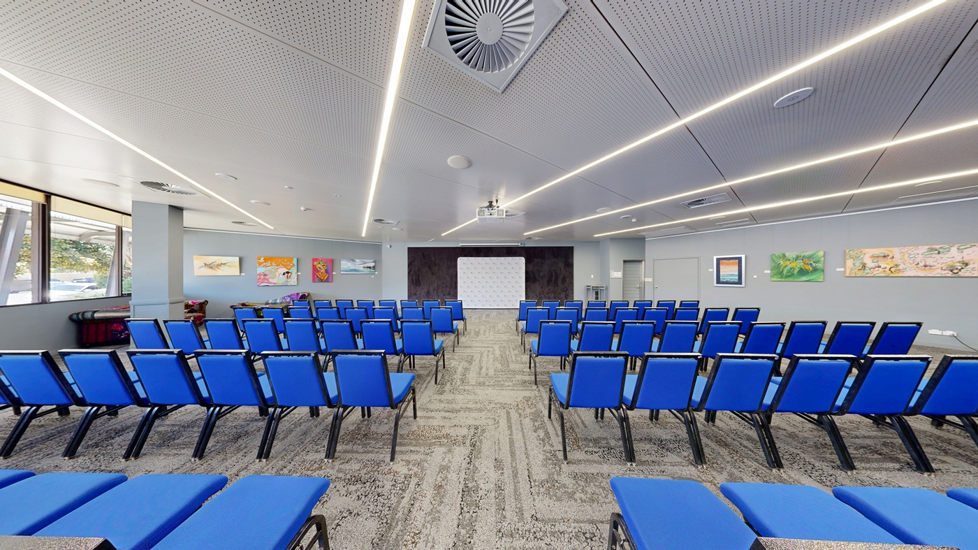 A conference room with rows of blue chairs facing a white projection screen at the front. The walls are adorned with colourful artwork. The ceiling features linear lights and a large air vent. The floor is carpeted with a patterned design. Windows on the left allow natural light to enter, and a small table with items is visible near the windows.
