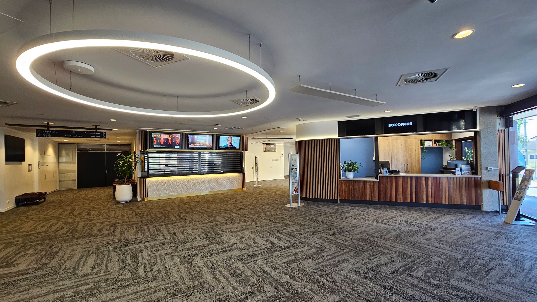 A spacious lobby with a patterned carpet in shades of green and grey. On the right, there's a box office with a wooden counter and a plant. In the centre, a circular light fixture hangs from the ceiling. To the left, a digital display board and a potted plant are visible. The ceiling has recessed lighting and air vents. Signs for different theatre rooms are visible in the background.