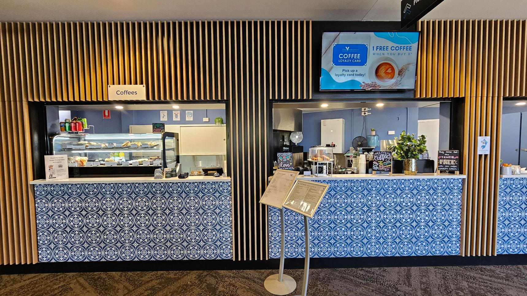 A café counter with two service windows, each adorned with blue and white patterned tiles. The left window displays a variety of snacks and drinks in a glass case, with a sign above reading "Coffees". The right window features a coffee machine and a small plant. Above, a digital screen advertises a coffee loyalty card. The background has vertical wooden slats, and a menu stand is positioned in front of the counter. The floor is carpeted with a geometric pattern.