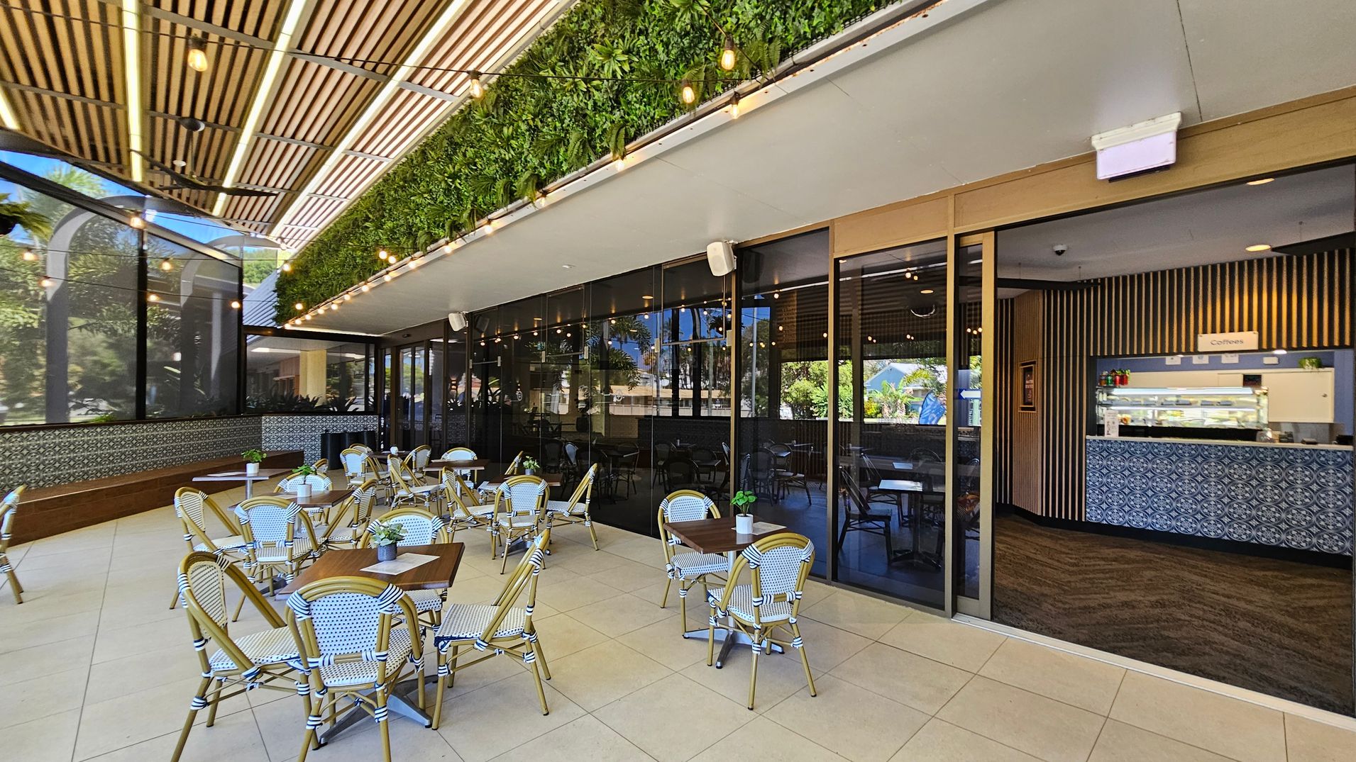 Outdoor café area with several tables and chairs on a tiled floor. On the left, a wooden bench runs along a wall with decorative tiles. The ceiling features wooden slats and string lights. A vertical garden with lush greenery is above the glass wall on the right, which separates the outdoor seating from the indoor café. Inside, a counter with patterned tiles displays a sign for coffee. Large windows reflect the surrounding greenery.