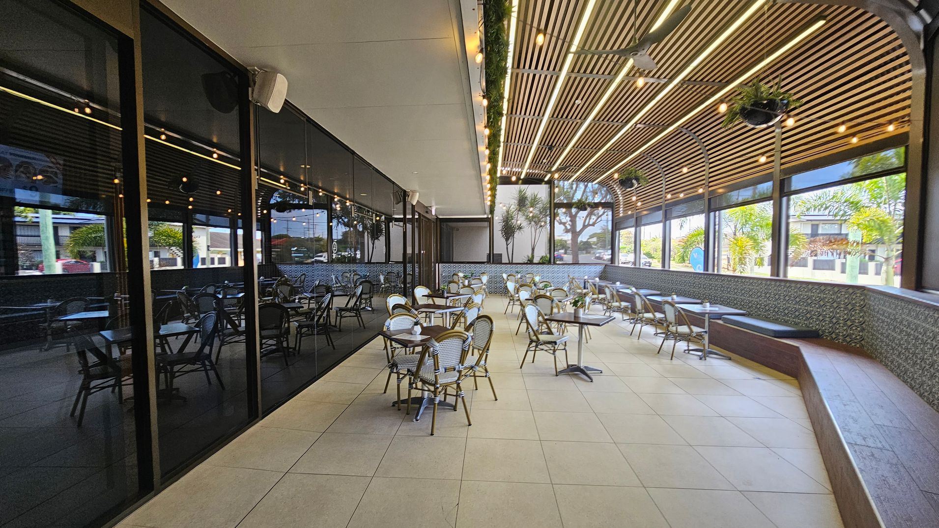 Outdoor seating area with numerous tables and chairs arranged on a tiled floor. On the left, a glass wall reflects the interior. The right side features a patterned wall with a long bench. The ceiling is wooden with hanging lights and potted plants. Large windows provide a view of trees and buildings outside.