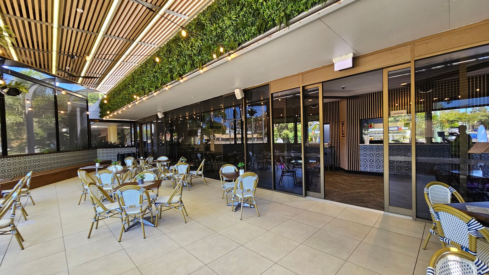 Outdoor café with wicker chairs and round tables on a tiled floor. The left side has a long bench with patterned tiles. The ceiling features wooden slats and hanging lights, with a green plant wall running along the top. Glass doors on the right lead to an indoor area with a counter and more seating. Reflections of trees and buildings are visible in the glass.