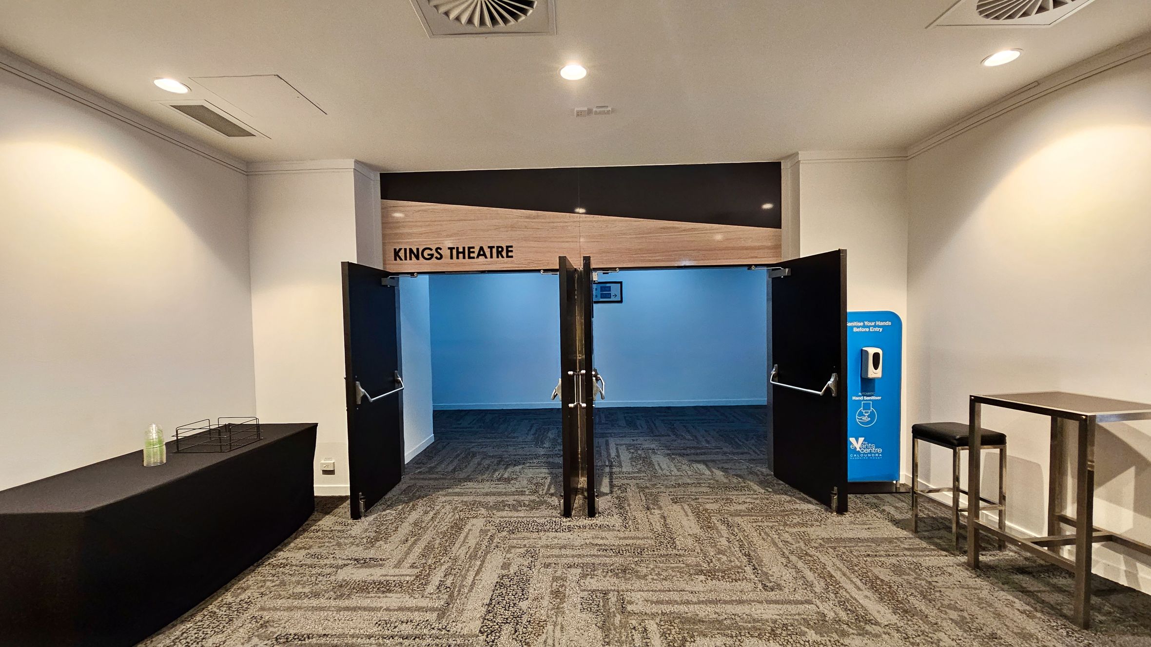 Entrance to Kings Theatre with open double doors on the left. The floor is carpeted with a geometric pattern. A small table with stools and a plant in a pot are nearby. On the right, a long bench is attached to the wall, which has several light fixtures. The ceiling is fitted with recessed lights and air vents. Further down the corridor, there are more plants and a digital display on the wall.