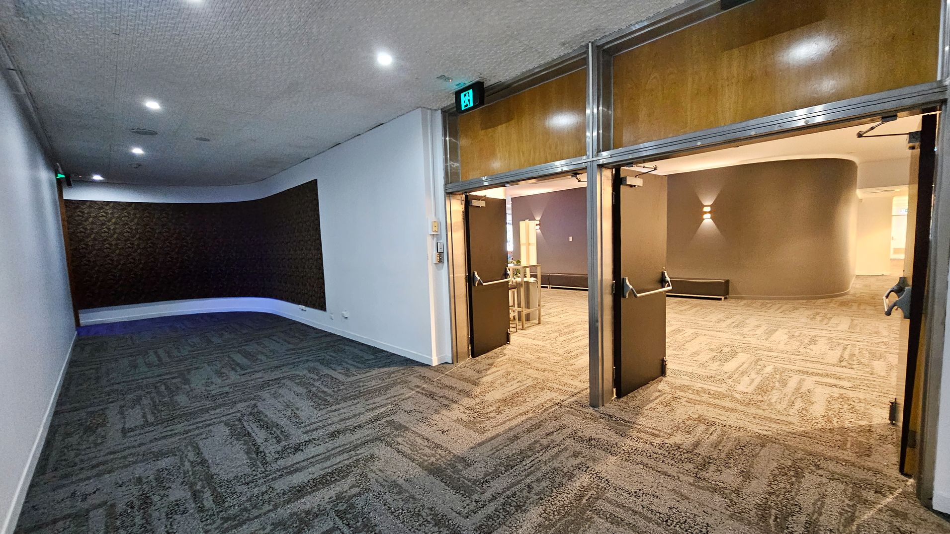 A hallway with patterned carpet flooring and a textured wall on the left. The ceiling is dotted with small lights. On the right, a set of double doors with wooden panels and metal frames is open, leading to another room with similar carpet and a curved wall. An illuminated exit sign is above the doors. The room beyond has a wall light fixture and some furniture, including a bench and a table.