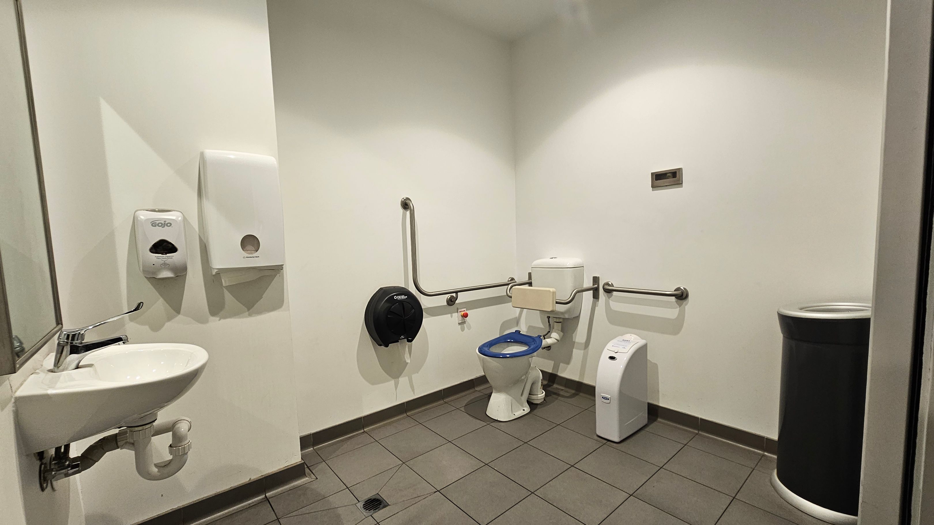 Accessible bathroom with a white sink and a mirror on the left wall. Above the sink are a soap dispenser and a paper towel dispenser. In the centre, a toilet with a blue seat is equipped with support rails on both sides. A black toilet paper dispenser is mounted on the wall beside it. On the right, a sanitary bin is placed against the wall. The floor is tiled in a light brown colour, and the walls are painted white.