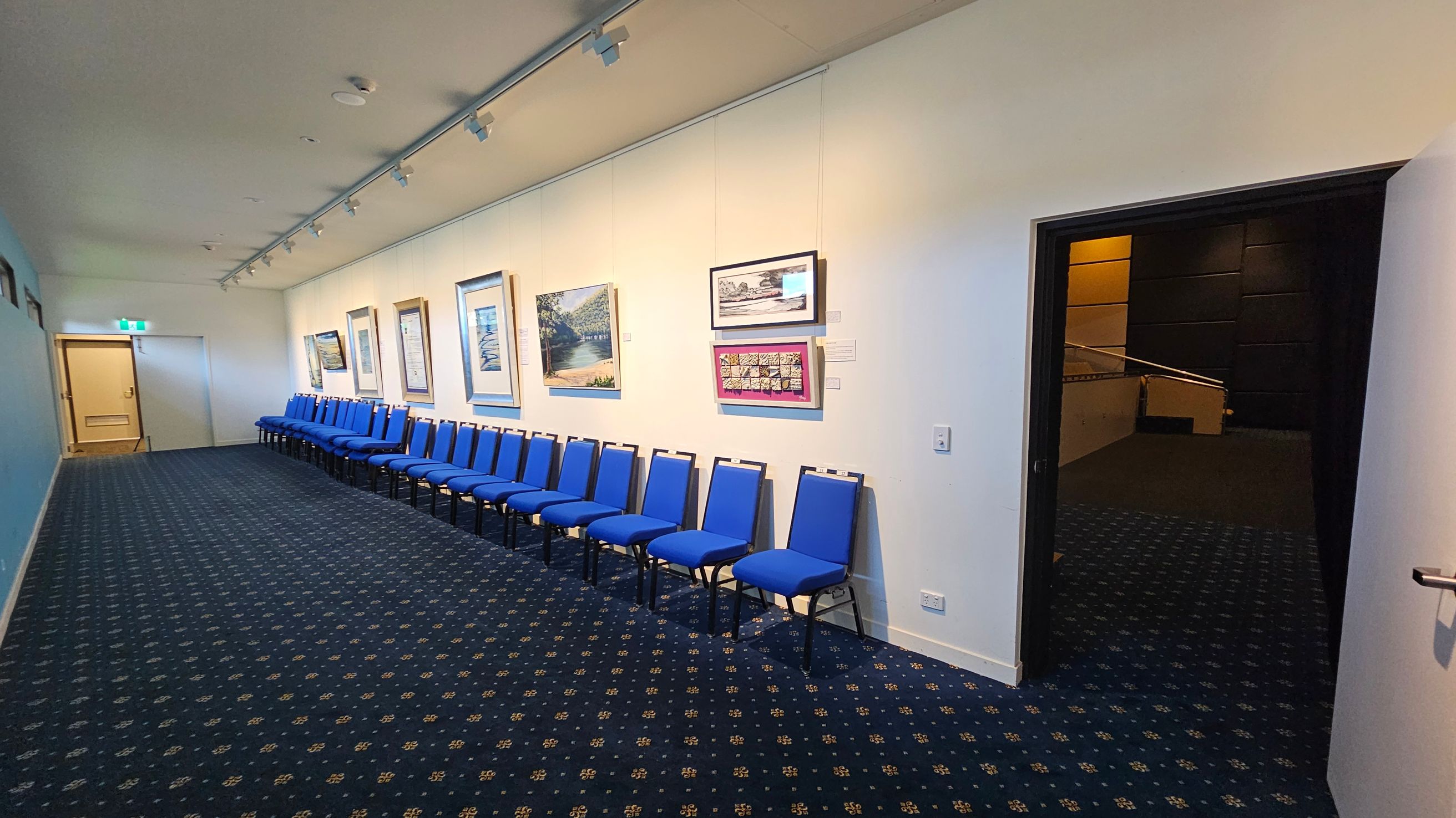 A hallway with a sign reading "Playhouse Theatre" above a set of carpeted stairs leading upwards. On the left wall, there is a sign with arrows indicating directions to the toilets and accessible facilities. On the right, a tall table with a black stool and a colourful cushion on the floor. The floor is carpeted with a patterned design, and the walls are painted in light colours.