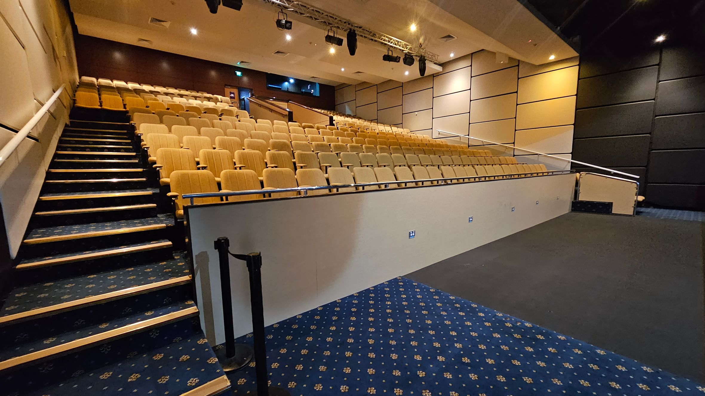 A lecture hall with tiered seating on the left, featuring rows of beige chairs. A staircase with a dark blue patterned carpet runs alongside the seats. The right side has a wall with beige and black acoustic panels. The floor in the foreground is a mix of dark blue patterned carpet and plain dark flooring. The ceiling is equipped with spotlights and a lighting rig.