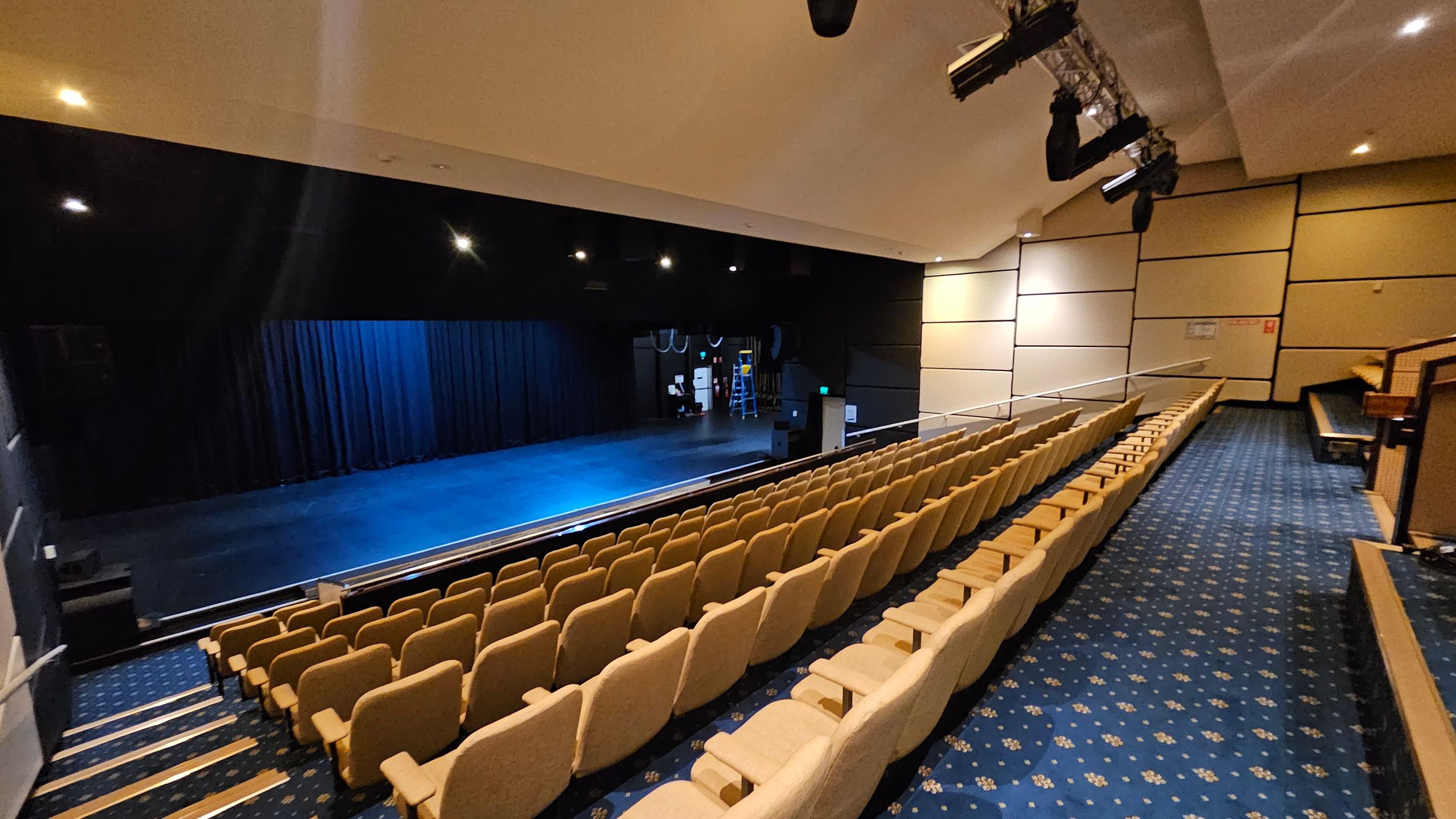 A theatre interior with rows of beige seats on the right, arranged in a tiered fashion. The floor is covered with a blue carpet featuring a floral pattern. On the left is a stage with a dark backdrop and blue lighting. Above the stage, several stage lights are mounted. The walls are lined with beige panels, and the ceiling is equipped with recessed lighting.