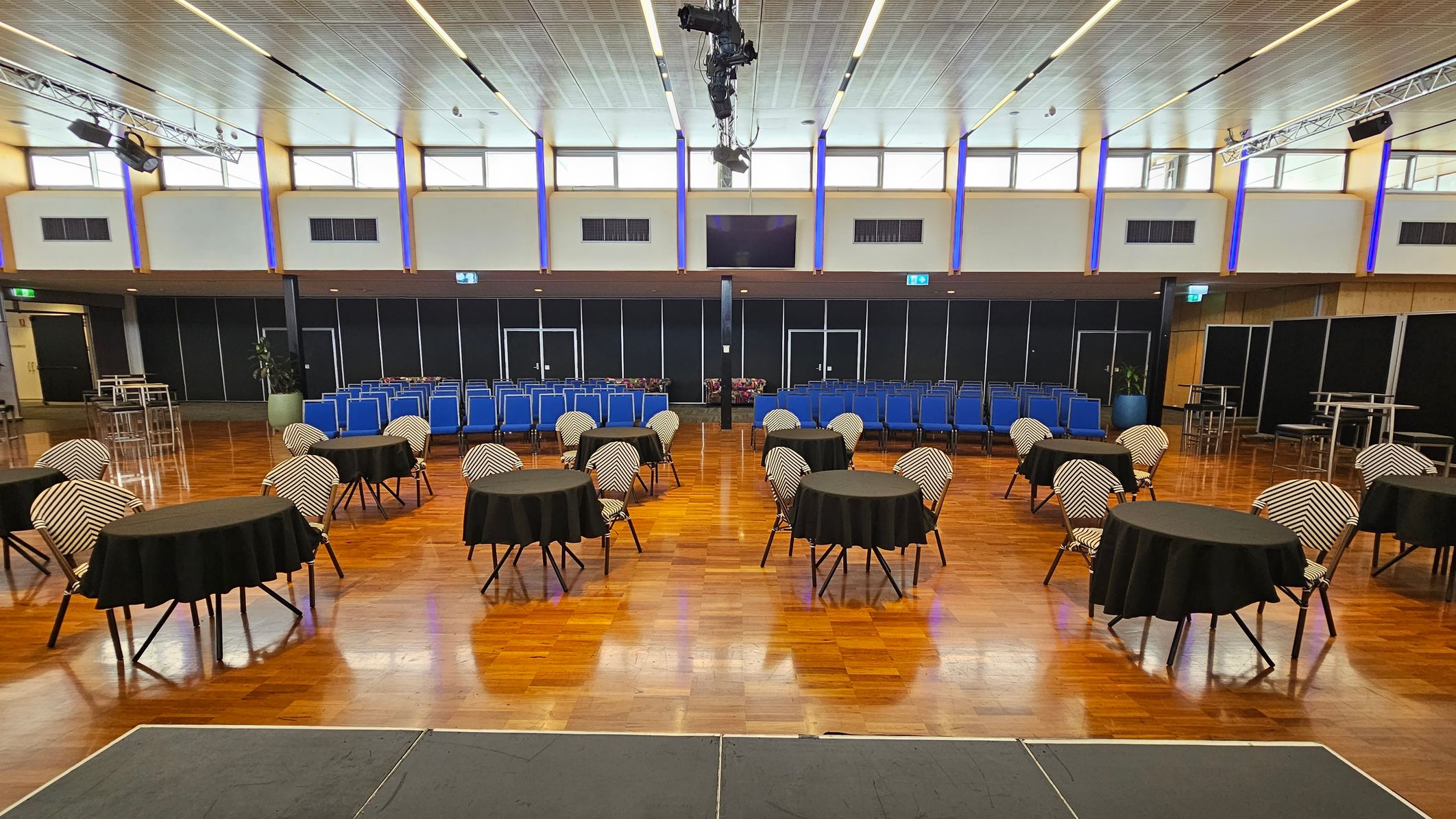 A spacious hall with a wooden floor and a high, panelled ceiling. On the left, there is a small stage and several round tables with black tablecloths and chairs. The right side features rows of blue chairs and a few tall tables with stools. The back wall has large windows and black panels, with lighting fixtures and speakers suspended from the ceiling.
