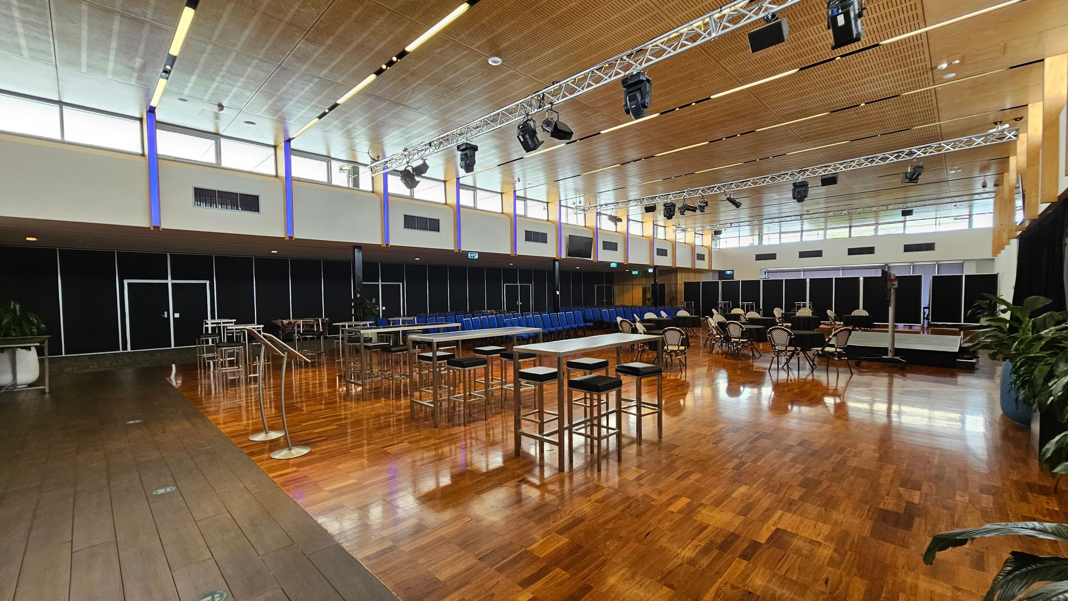 A spacious hall with a polished wooden floor. In the foreground, several round tables are covered with black tablecloths, surrounded by chairs with black and white striped covers. In the background, rows of blue chairs are neatly arranged. The ceiling is high with a grid pattern and features lighting fixtures. The walls have large windows near the ceiling, allowing natural light to enter. On the sides, there are tall plants in pots and some high tables with stools.