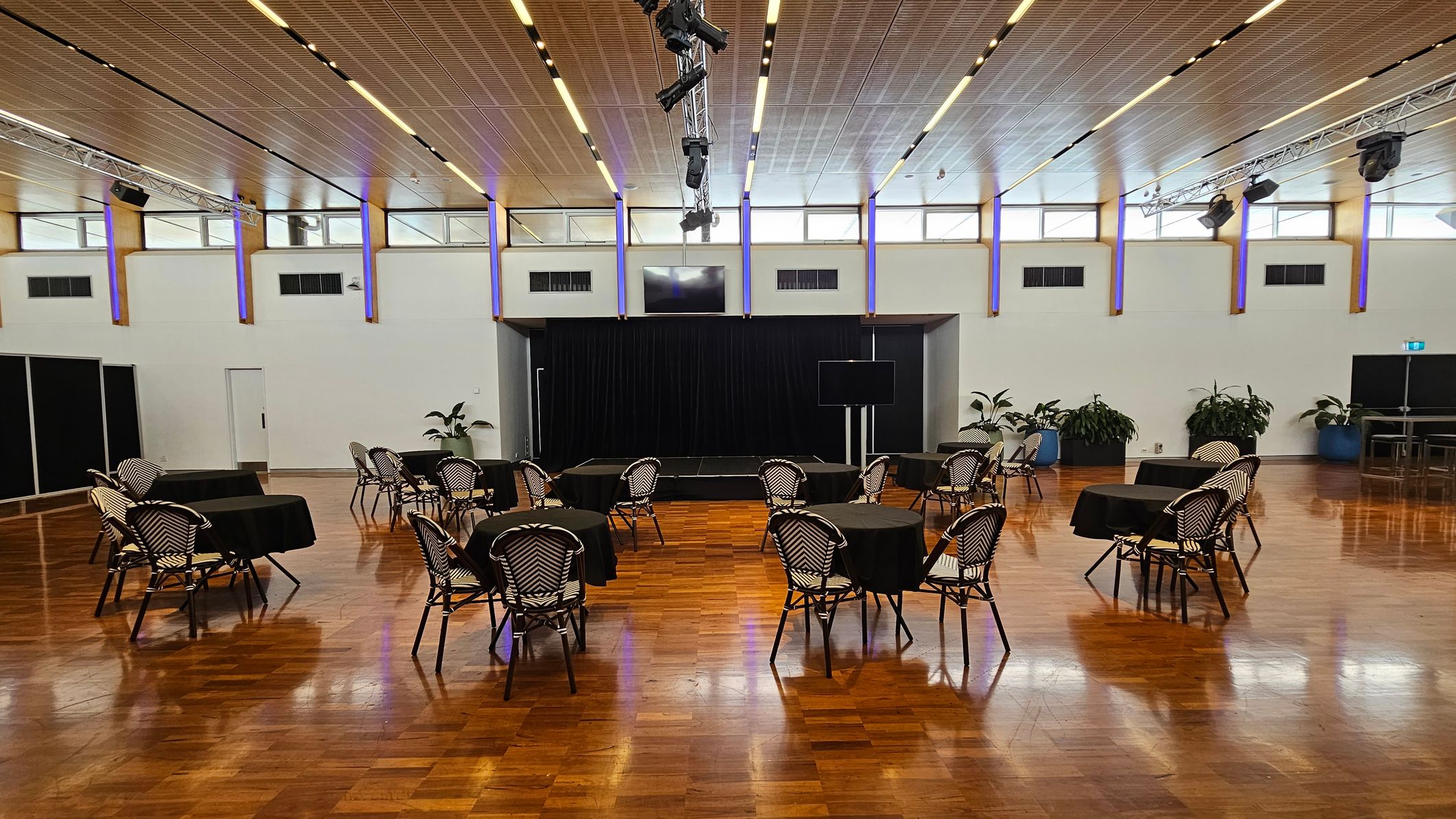 A spacious room with a polished wooden floor and a high ceiling featuring recessed lighting. In the centre, there are several round tables covered with black tablecloths, each surrounded by chairs with a woven pattern. At the back, a small stage is set against a black curtain. Potted plants are placed along the walls, and a large screen is visible to the right. The walls have high windows, allowing natural light to enter.