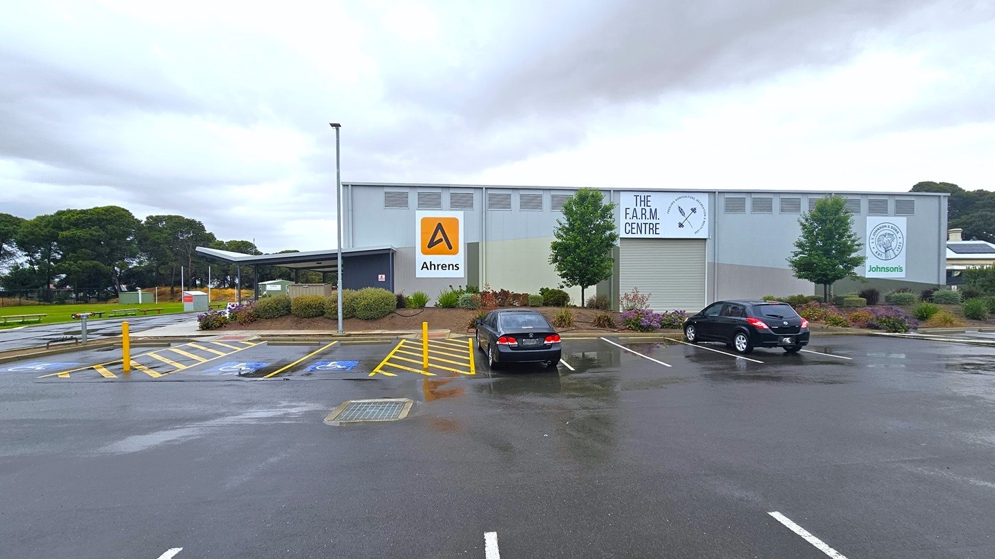 The car park is located near the building. Accessible parking is marked with blue wheelchairs, yellow diagonal lines, and bollards. Two black cars are parked. Behind the car park is a grey industrial-type building with the sign "The Farm Centre." Advert signs are displayed on the building wall. The building is surrounded by flowers, ornamental landscapes, and trees. The left section features a grassy area.