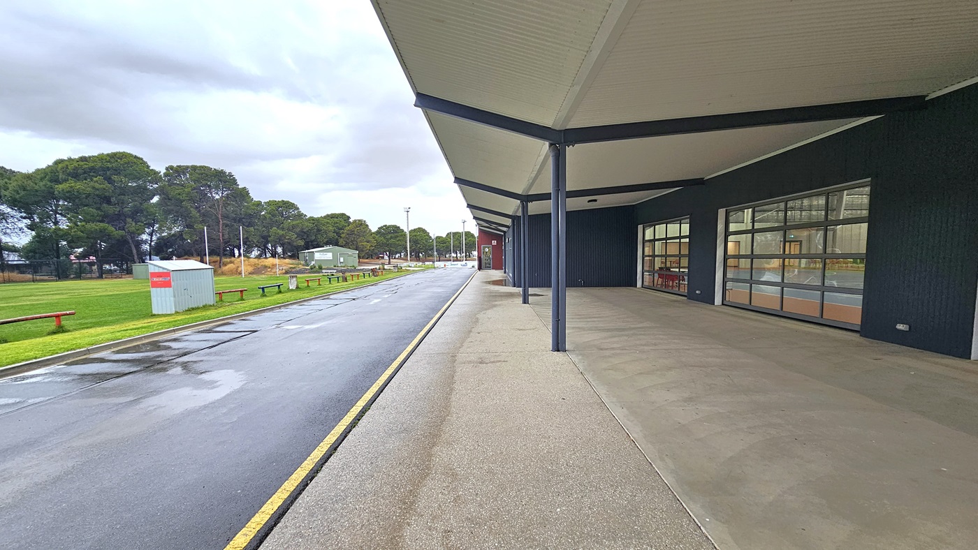 On the left is a grassy area with bench seating. The road separates the grassy area from the building on the right. The front of the building features a pitched white ceiling supported by large metal beams. On the right is a dark grey wall with large window glass.