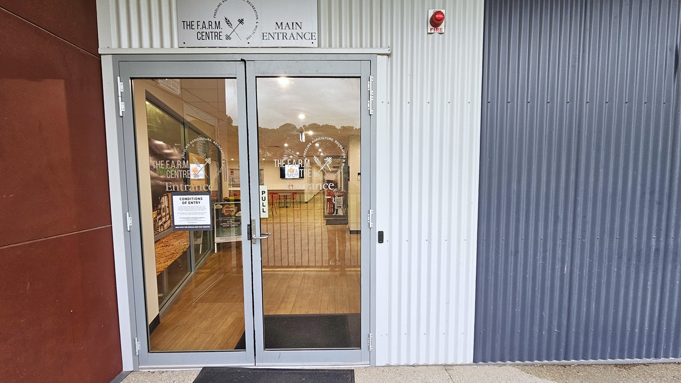 Entrance to the building is through a manual outward-opening door equipped with a non-slippery mat. Facility signage is placed on the wall. Posters and conditions of entry are affixed to the glass door along with stickers bearing the facility name. To the left is a concrete wall painted in brown, while on the right is a wall with corrugated design in dark grey. A fire safety light is placed on top.
