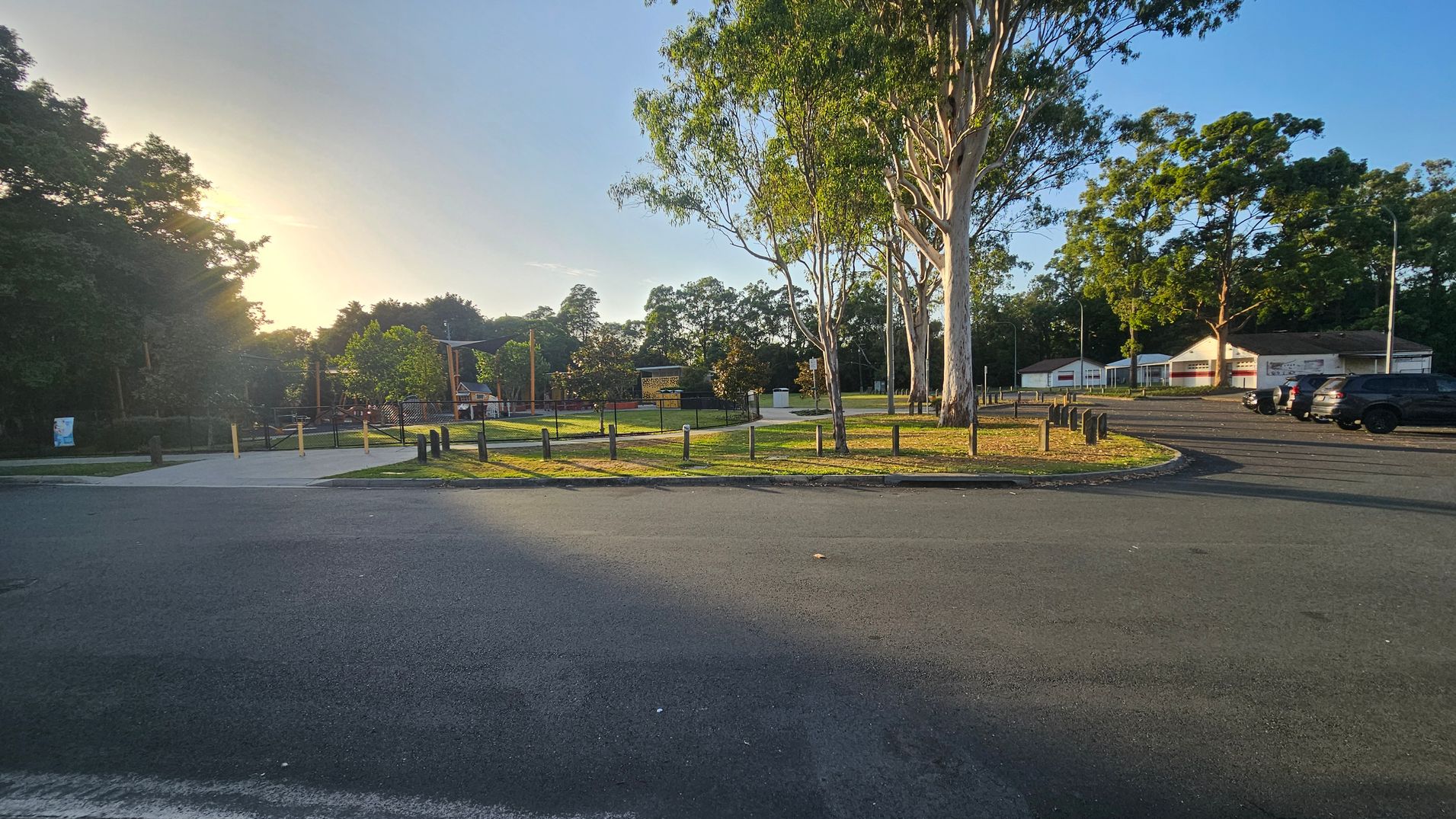 A park with a paved path and grassy area in the centre, surrounded by tall trees. On the right, there are several parked cars and a few buildings with white walls and red signage. The left side has more trees and a fenced area. The ground is a mix of asphalt and grass, and the sun is setting in the background, casting long shadows.