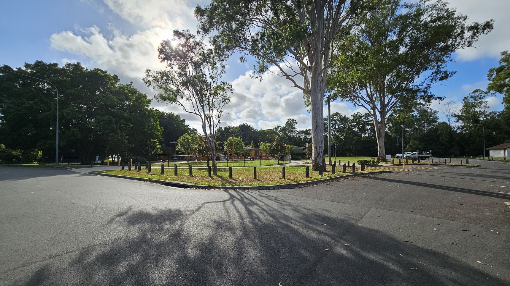 A park with a grassy area in the centre, surrounded by a low wooden barrier. Tall trees with sparse leaves are scattered throughout, casting shadows on the ground. To the right, a paved parking area with a few cars is visible. On the left, dense foliage forms a backdrop. The sky is partly cloudy, and the sun is partially obscured by the trees. The road in the foreground is smooth and dark, with tree shadows stretching across it.