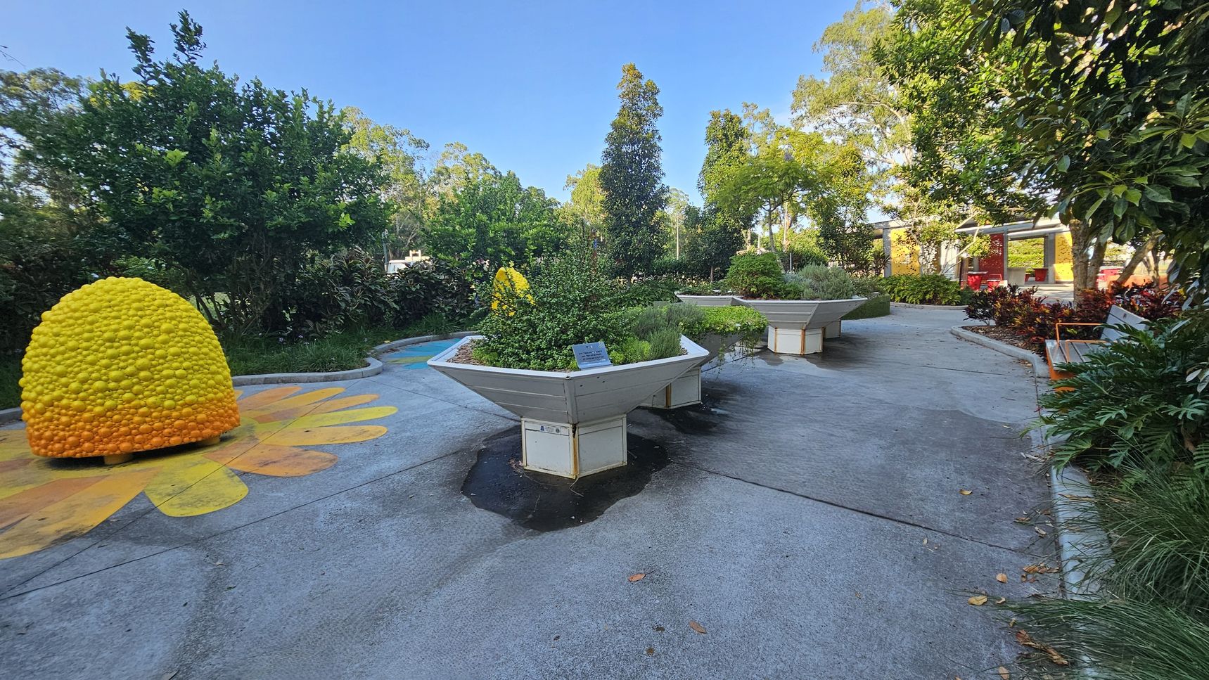 A garden area with a concrete path. On the left, there's a large, dome-shaped sculpture resembling a beehive, painted in yellow and orange. In the centre, there are large, hexagonal planters filled with various green plants. To the right, a wooden bench is surrounded by lush foliage. In the background, trees and shrubs create a dense, green backdrop, and a structure with red accents is visible. The ground is decorated with colourful patterns.