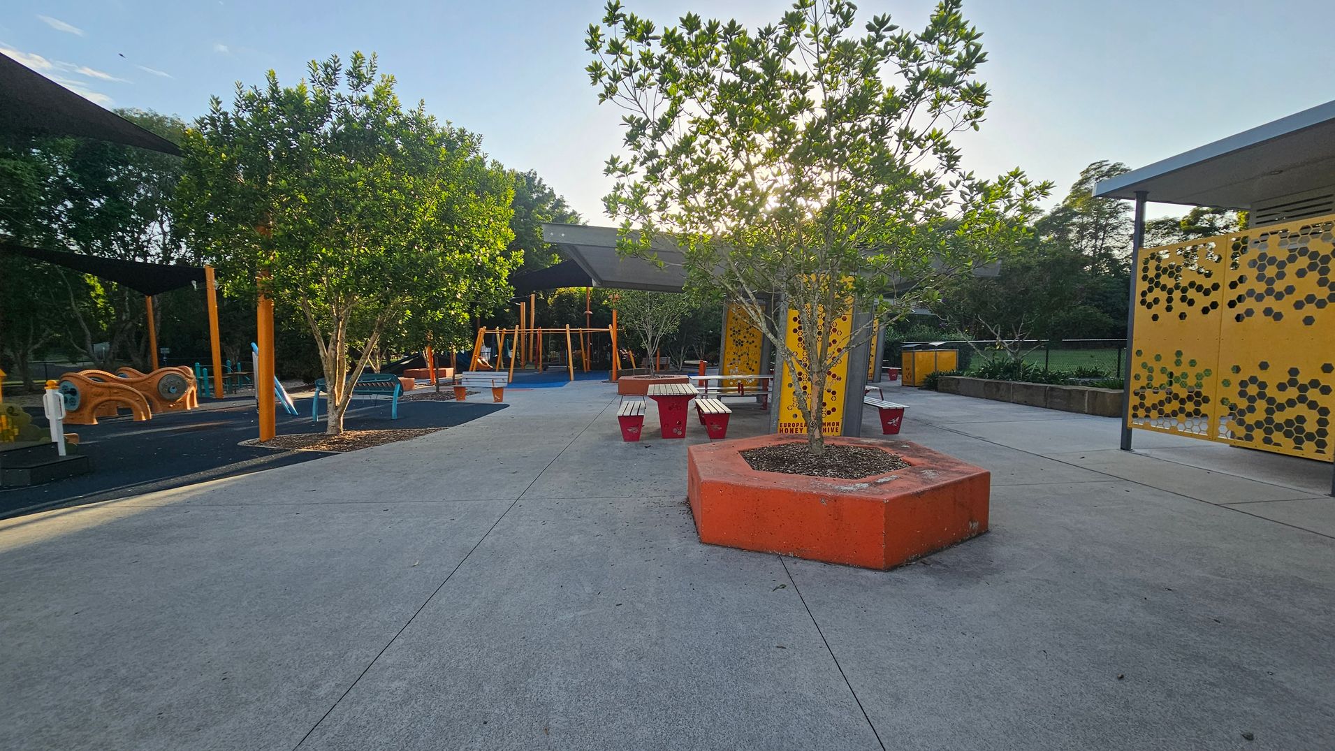 A playground with a concrete path in the foreground. On the left, there are trees and playground equipment with orange and blue structures. In the centre, a tree is planted in an orange hexagonal planter, surrounded by benches and red stools. On the right, a yellow panel with a honeycomb pattern is visible, along with more trees and a grassy area in the background. The area is shaded by large canopies.