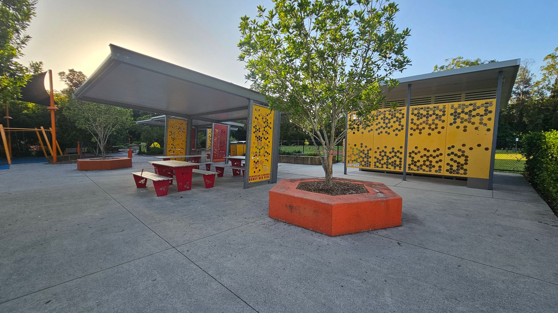 A playground area with a concrete floor. On the left, there's a play structure with orange supports. In the centre, a covered seating area with red benches and tables is visible. A tree is planted in an orange hexagonal planter. On the right, a structure with yellow panels featuring a honeycomb pattern. In the background, there are trees and a grassy area enclosed by a fence.