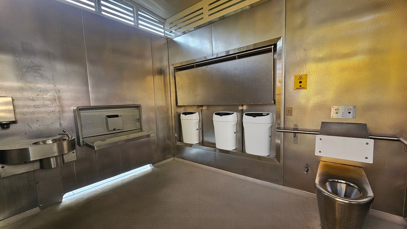 A public restroom with metallic walls and a concrete floor. On the left, a stainless steel sink with a soap dispenser is mounted on the wall. In the centre, a baby changing station is above three white bins. On the right, a stainless steel toilet is visible, with a sharps disposal box mounted above it. The ceiling has a vented design, allowing light to filter through.