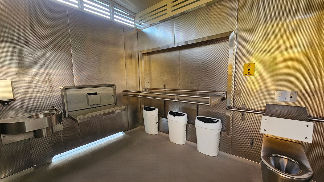 A public restroom with metallic walls and a concrete floor. On the left, there's a stainless steel sink with a soap dispenser above it. Next to the sink is a baby changing station. In the centre, three white bins are mounted on the wall. On the right, a stainless steel toilet with a safety rail and a small control panel above it. A yellow sharps disposal sign is visible on the wall. The ceiling has a vented section allowing light to filter through.