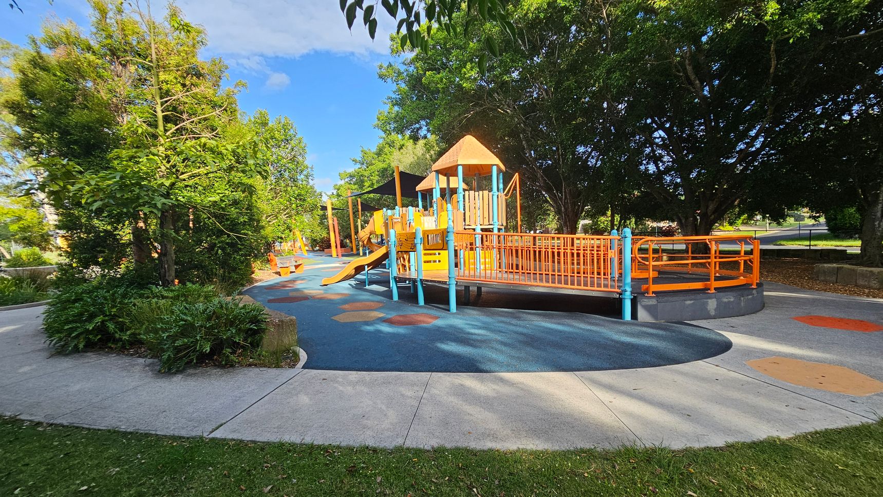 A playground with a colourful play structure featuring orange and blue elements, including slides and ramps, is in the centre. The ground is covered with a blue and orange rubber surface. On the left, there are lush green trees and shrubs. In the background, more trees provide shade. The foreground has a concrete path and grass.