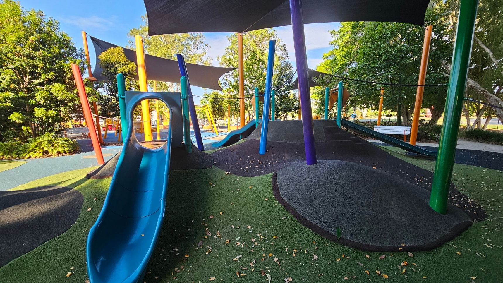 A playground with a blue slide on the left, leading down to a surface covered in green and black rubber. Colourful poles in blue, orange, and green support overhead shade sails. In the background, there are trees and additional playground equipment, including swings. A white bench is visible to the right, surrounded by more trees and foliage. The ground is scattered with fallen leaves.