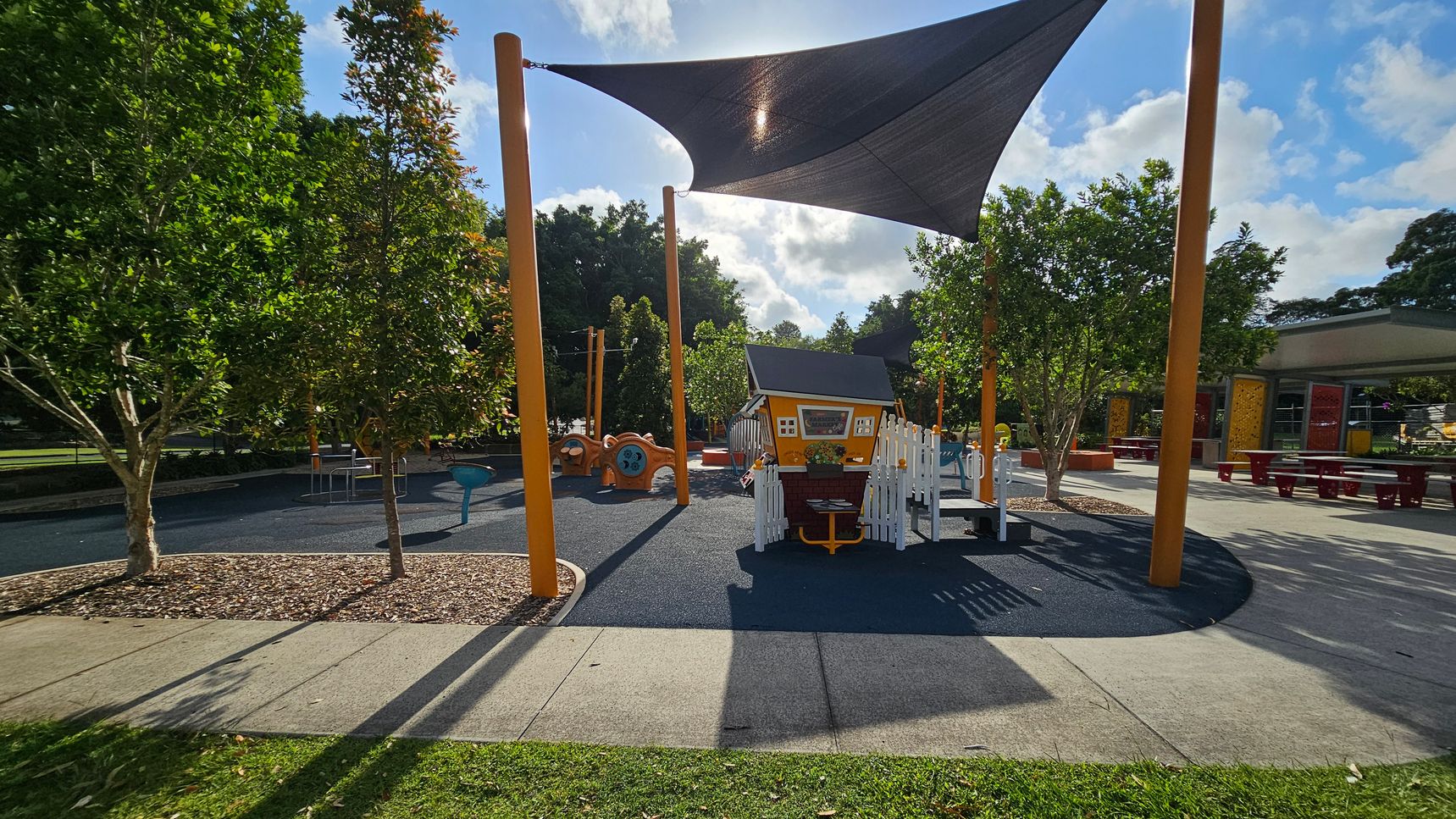 A playground with a small playhouse in the centre, surrounded by a white picket fence. The ground is covered with a soft, dark surface. Tall orange poles support a large triangular shade sail overhead. To the left, there are trees and a play structure with a blue seat. On the right, picnic tables are visible under a shelter. The foreground has a grassy area and a concrete path, while more trees and playground equipment are in the background.