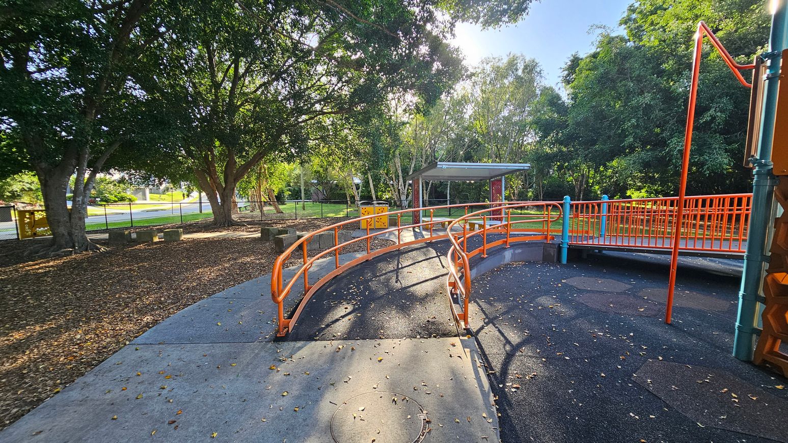 A playground with an orange railing bridge in the centre, leading to a covered area in the background. The ground is a mix of concrete and rubberised surface with scattered leaves. On the left, large trees provide shade, and a fence runs along the perimeter. On the right, more playground equipment is partially visible. The area is surrounded by greenery, creating a natural setting.