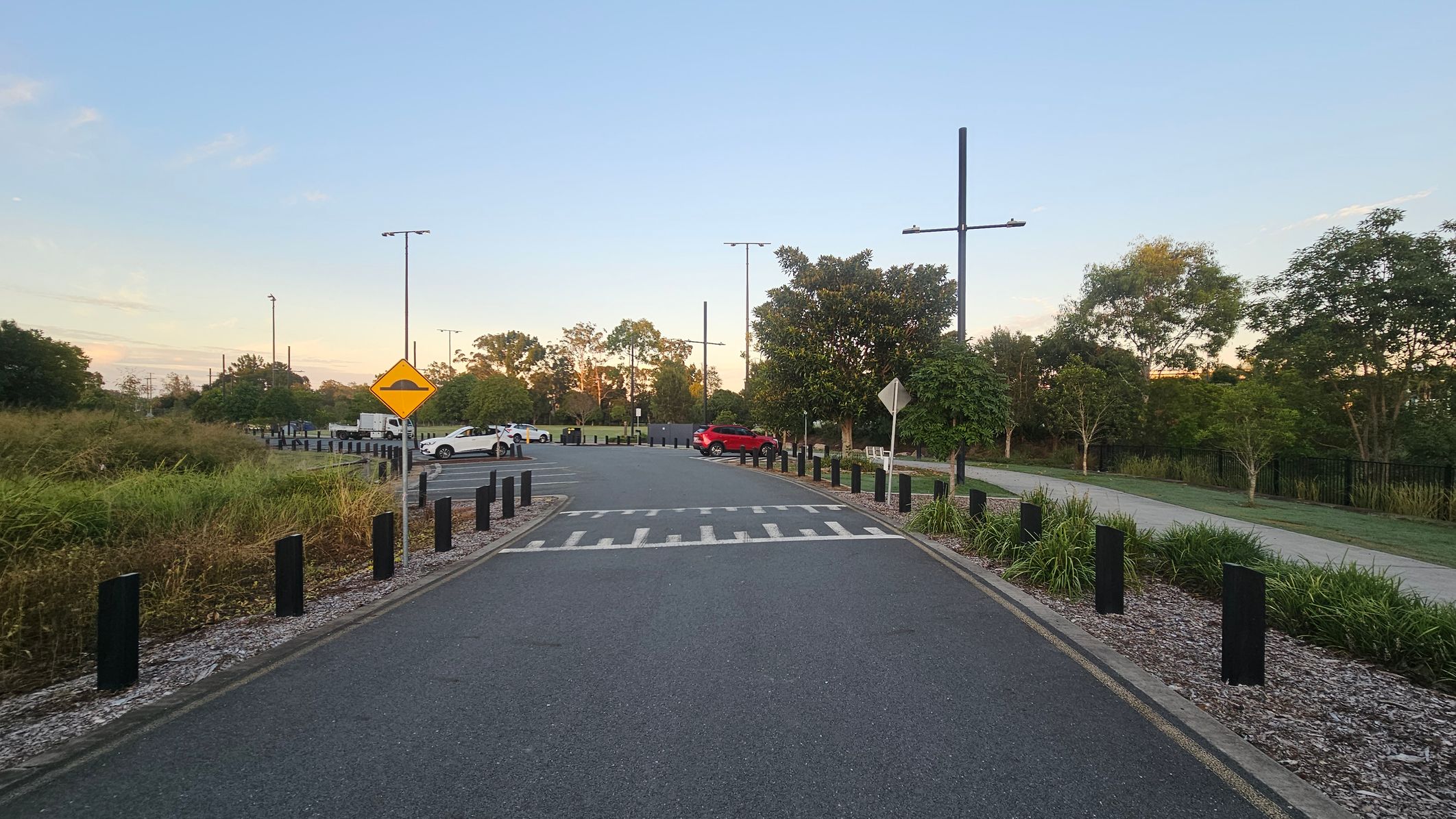 A road with a speed bump sign in the centre, flanked by black posts and gravel on both sides. On the left, there's tall grass and a few parked cars, including a white one. On the right, a red car is parked near a path lined with trees and shrubs. Tall streetlights are visible along the road, and trees are in the background.