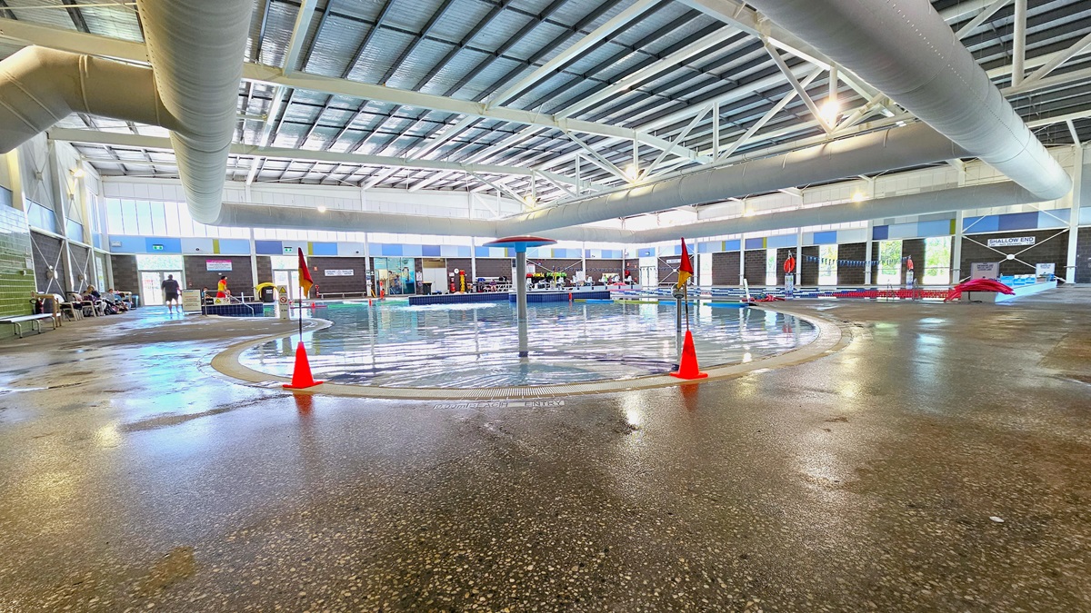 Access to the Aquatic Centre is through manually operated outward-opening glass doors. The centre has a high ceiling with large air-conditioning vents, metal beams, and bright lights. On the right, there is a large banner, while round yellow-green signages are affixed to the brick wall on the left. Additional informational posters are on the glass door. The pool area is visible from here.